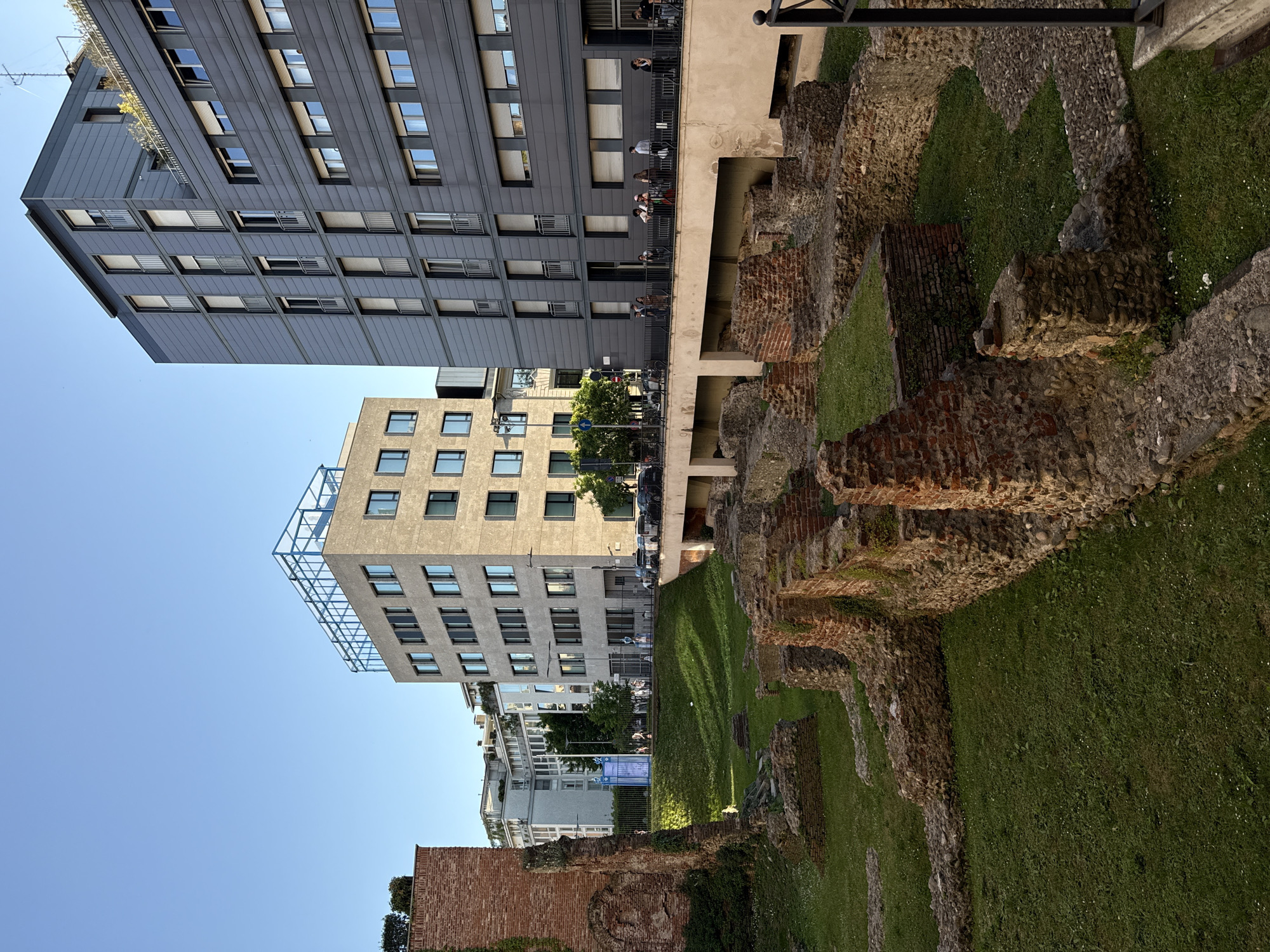 The image shows a street scene in Milan, Italy, featuring a mix of modern architecture and ancient Roman ruins. On the left are two contemporary buildings with gray facades and symmetrical window arrangements, suggesting residential or office use. Adjacent to these buildings are the remains of an ancient Roman wall constructed from bricks and stones, with green grass growing around the ruins. People can be seen standing on a balcony, overlooking the ancient ruins, which indicates that the site is accessible and possibly a tourist attraction. The sky is clear and blue.
