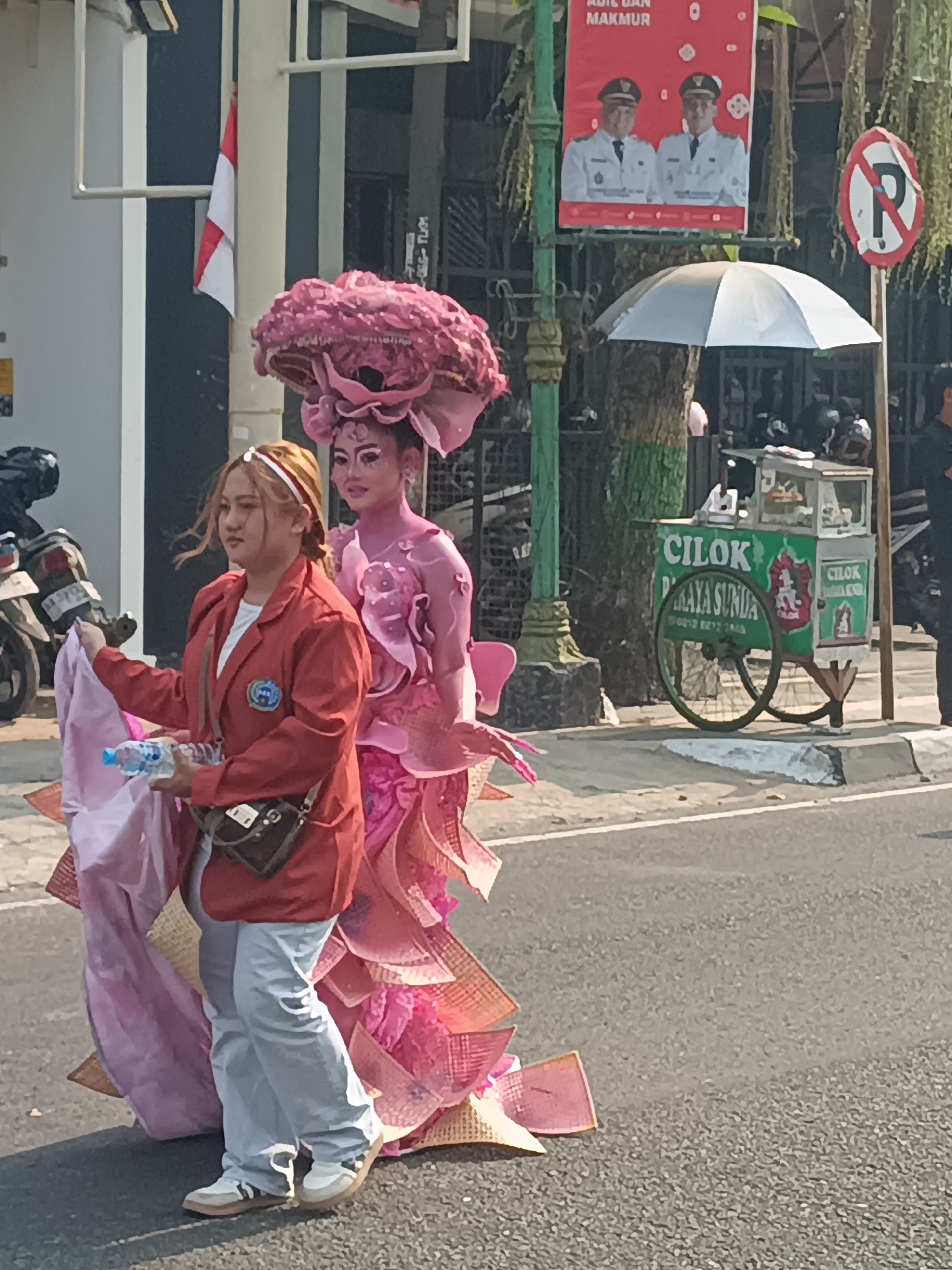 Two women are walking on a street in Sleman, Indonesia. The woman on the left is wearing a red jacket, white t-shirt, light blue pants, and white sneakers. She is holding a pink fabric and a plastic bottle of water. The woman on the right is wearing a large pink floral headpiece and a pink costume with elaborate ruffled details. She has full face makeup, including dramatic eye makeup and pink body paint. In the background, there is a food cart labeled "CILOK PRAYA SUNDA," a white umbrella, a no-parking sign, and a poster featuring two men in uniform. A motorcycle is visible on the left side of the image. The scene appears to be a parade or festival.