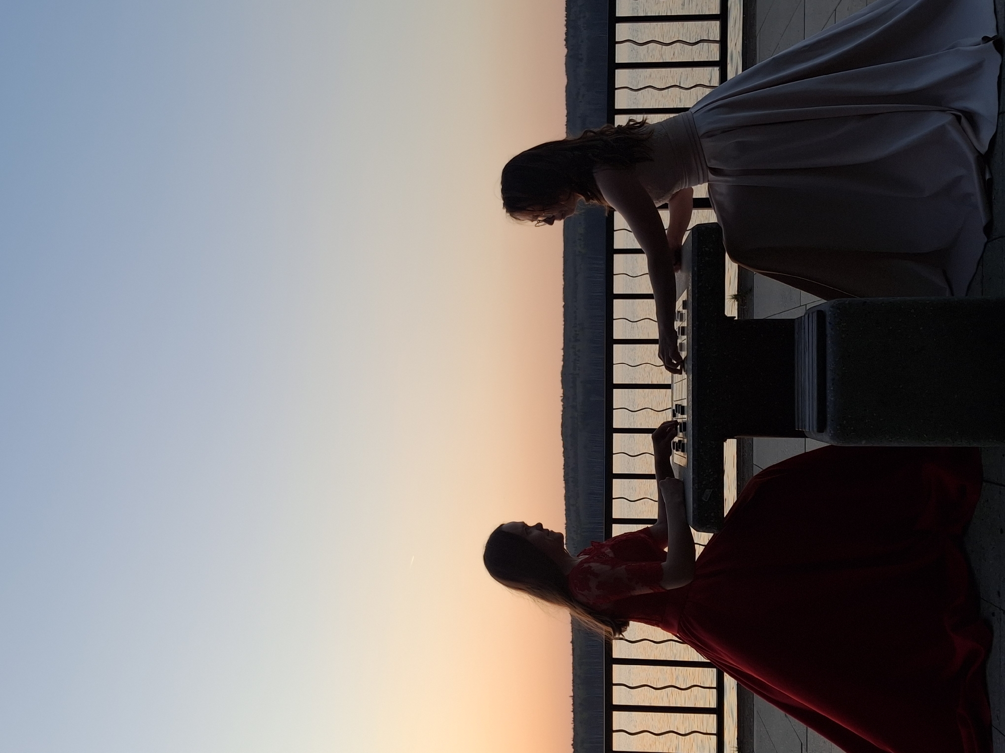 Two women in formal gowns stand facing each other over what appears to be an outdoor piano keyboard. The woman on the upper right is wearing a light-colored gown, and the woman on the bottom left is wearing a red gown. They are near a railing with the suggestion of water visible behind it. The sky is a gradient of blue to orange, indicating either sunrise or sunset. The image orientation is rotated; the horizon appears on the left. The scene suggests a promenade area with decorative elements. Based on the style of dress, architectural features, and the appearance of the landscape, the location is potentially the city of Chaikovsky, Russia.