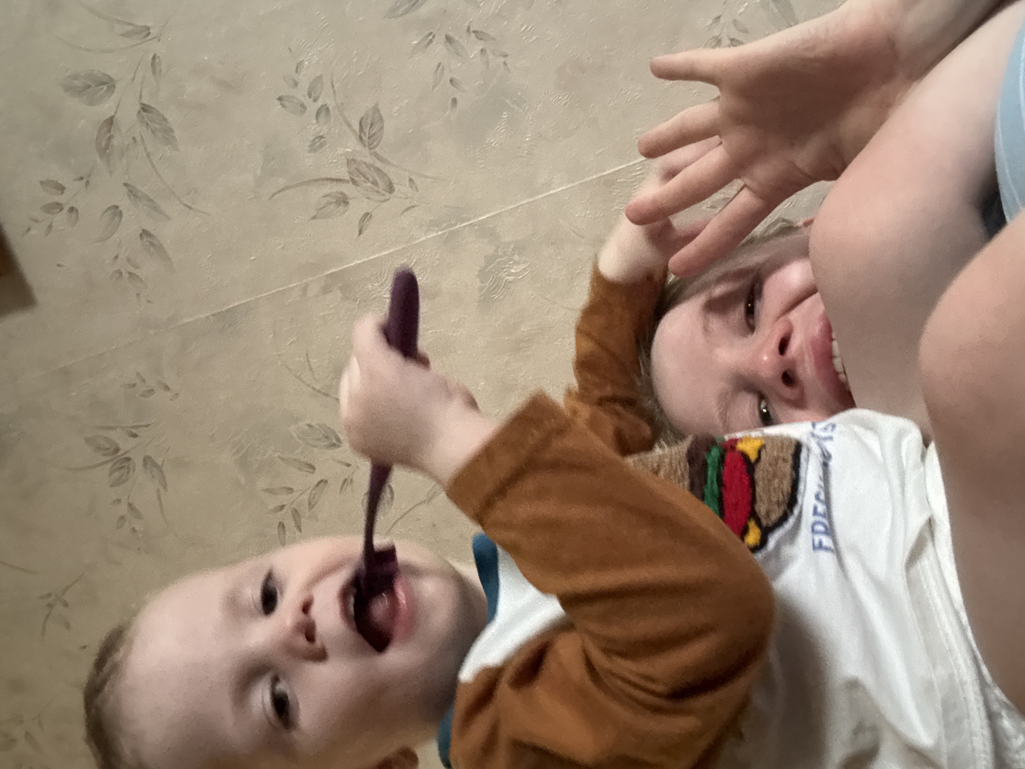 This candid, overhead shot captures two children engaged in playful interaction, likely indoors in Saint Petersburg, Russia. The perspective looks down onto the children, who appear to be lying on a surface against a backdrop of light-colored wallpaper adorned with a subtle, greyish floral or leaf pattern.
In the foreground and to the left, a young child, possibly a toddler, with light hair and fair skin, is seen. They are wearing a brown long-sleeved top over a white garment, with a hint of blue fabric near the shoulder. This child has their mouth wide open, possibly mid-laugh or attempting to eat, while holding a purple, spoon-like utensil or toy towards their mouth. Their expression is lively and engaged.
To the right and slightly behind the younger child, an older child or teenager is partially visible. This individual also has light hair and is smiling broadly, looking up towards the camera. Their left hand is raised, fingers splayed, as if in a wave or playful gesture to block the camera. They are wearing a white shirt with a colorful embroidered patch on the chest, resembling a layered burger design (red, green, brown, yellow). Blue text, partially visible as "FDEC", is printed vertically on the white shirt. A light blue garment, possibly shorts or pajamas, is seen around their lower torso/legs.
The overall mood of the image is joyful and spontaneous, capturing a moment of children's play. The time of day is not discernible, but the indoor setting suggests a casual, home environment.