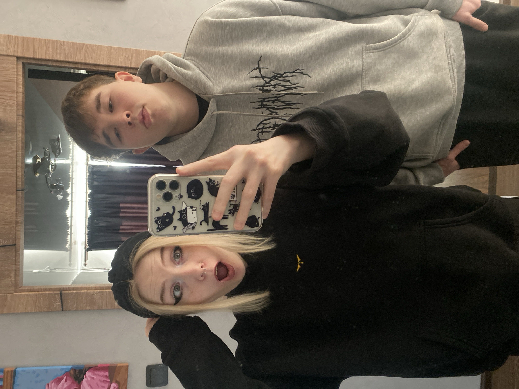 Two individuals are taking a selfie in front of a mirror. The person in the foreground wears a black hoodie and a black hat, has blonde hair, and is making an exaggerated surprised expression with their mouth open. The person in the background wears a grey hoodie with a dark design and is making a pursed-lip expression. The phone is visible with a clear case decorated with cats. The background includes a wooden-framed mirror reflecting a room with curtains and a painting. The location and its city or country are not discernible from the image.