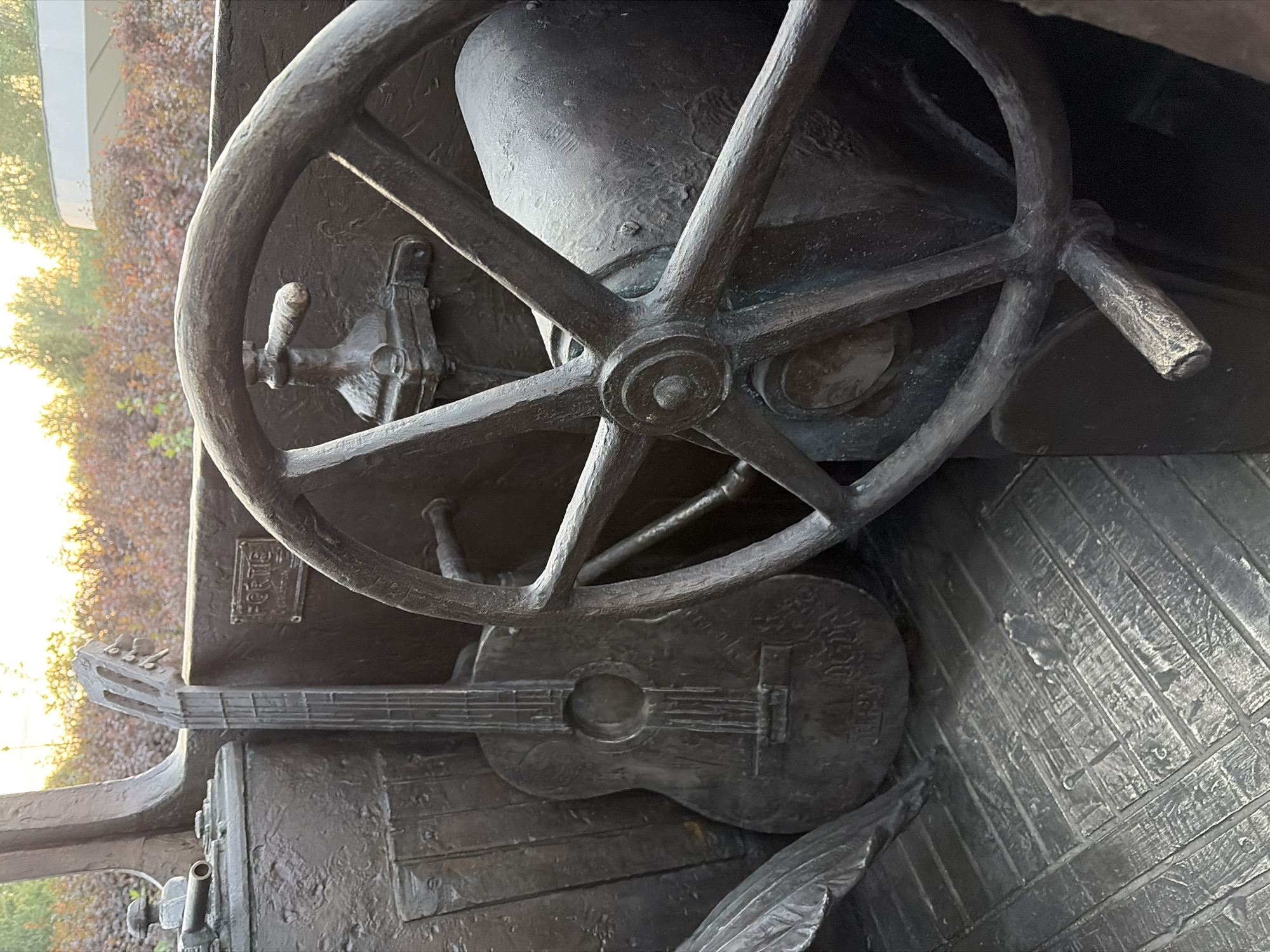 This image depicts a bronze sculpture of a vintage vehicle's interior, featuring a steering wheel, a guitar, and what appears to be a dashboard. The sculpture is located in Yekaterinburg, Russia. The steering wheel is large and spoked, with a lever mechanism visible. To the left of the steering wheel, a guitar is positioned upright, its body and neck clearly rendered in bronze. The dashboard area shows textured surfaces and a rectangular plate with the inscription "FORTIS." The floor of the sculpture is textured to resemble wooden planks. In the background, out-of-focus foliage and trees are visible, with a hint of a building and the sky at sunset. The overall impression is of a static, artistic representation rather than a live scene.