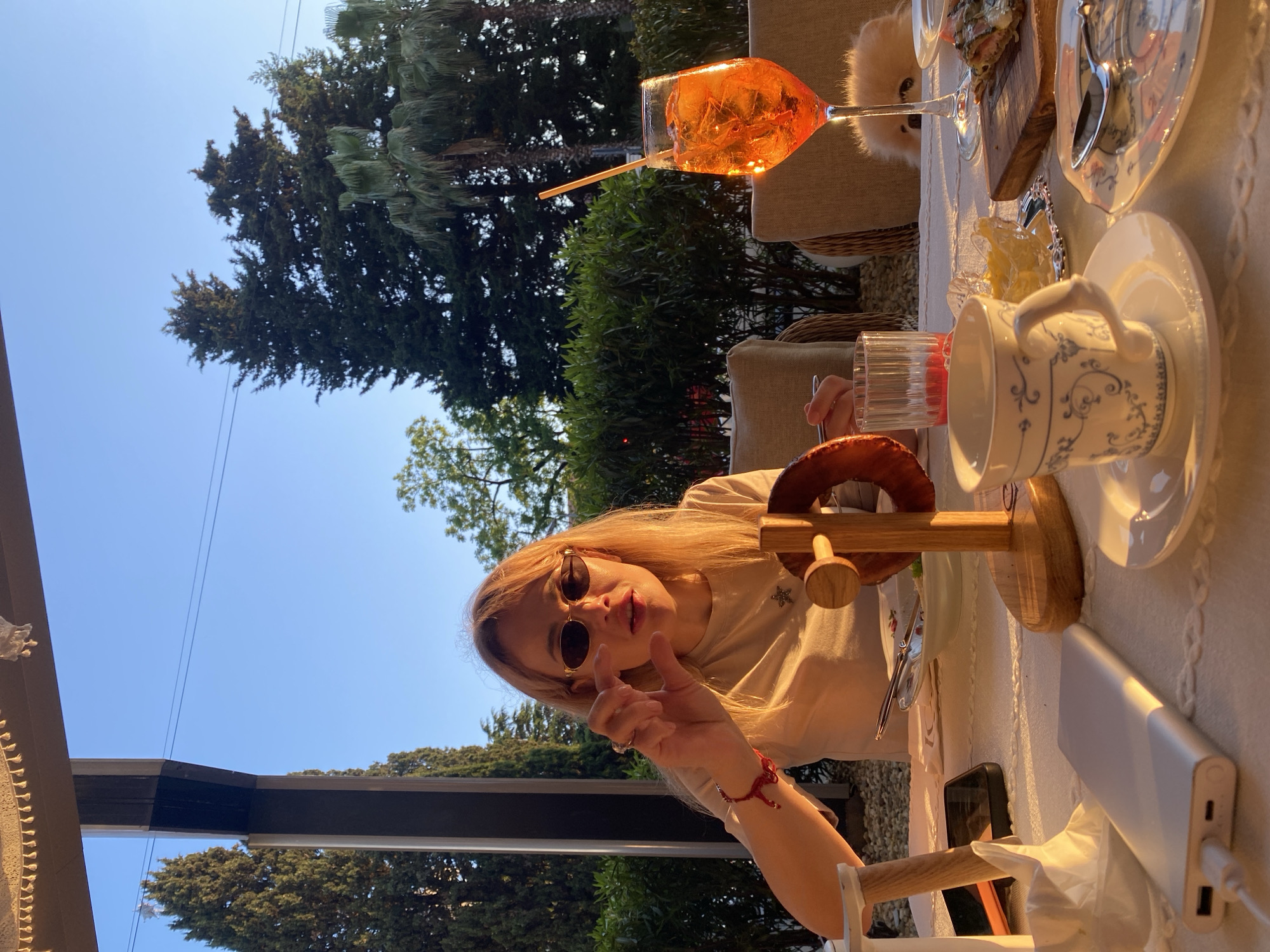 An outdoor dining scene in Sochi, Russia, during the golden hour, featuring a woman, a dog, and various food and drink items on a white-clothed table. The woman, with long blonde hair and wearing dark oval sunglasses, a beige t-shirt with a star emblem, and a red bracelet on her left wrist, is seated at the table, facing the camera with her mouth slightly open and her left hand making a small gesturing motion. On the table, from left to right, are a silver power bank plugged into a white cable, a crumpled white paper napkin, a black smartphone, a partially consumed plate of food, a wooden stand holding a large pretzel-shaped item, a glass of red liquid, a white teacup with blue patterns on a saucer, a small silver dish containing a yellow substance, and a patterned plate with a spoon. Further to the right, a wooden cutting board holds a savory dish, and a large stemmed glass contains an orange Aperol Spritz cocktail with ice and a straw. A small, light-colored fluffy dog is visible peeking from behind the Aperol Spritz glass. The background shows a clear blue sky, numerous tall green trees including conifers and palm trees, and lush green bushes, indicative of a southern climate. Dark architectural pillars and a light-colored ceiling with decorative fringe suggest a covered patio or veranda. Power lines are visible against the sky. The warm, directional sunlight casts long shadows across the table. Another person's arm with cutlery is partially visible to the left.