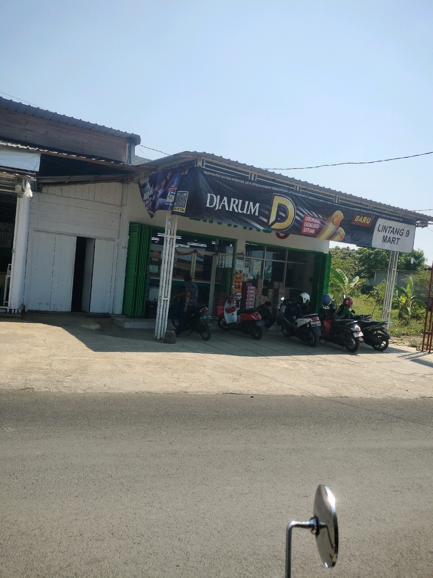 This image captures the exterior of a small shop named "LINTANG 9 MART" in Tersono, Indonesia, on a bright, sunny day. The shop, a single-story building with a white facade and green shutters, has a prominent banner across its front advertising "DJARUM" cigarettes. The banner also features an image of cigarettes and text indicating they are "DIBUNGKUS EKSKLUSIF SATUAN" (packaged exclusively per unit) and "BARU" (new). In front of the shop, several motorcycles are parked along the sidewalk, with one person seated on a dark motorcycle. The shop's entrance consists of a white door on the left and green collapsible doors on the right, leading into what appears to be a retail space. The ground in front of the shop is paved, and beyond it, a road is visible. In the foreground, a motorcycle's side mirror is partially captured in the frame. The background shows lush green trees and vegetation, suggesting a rural or semi-urban setting.