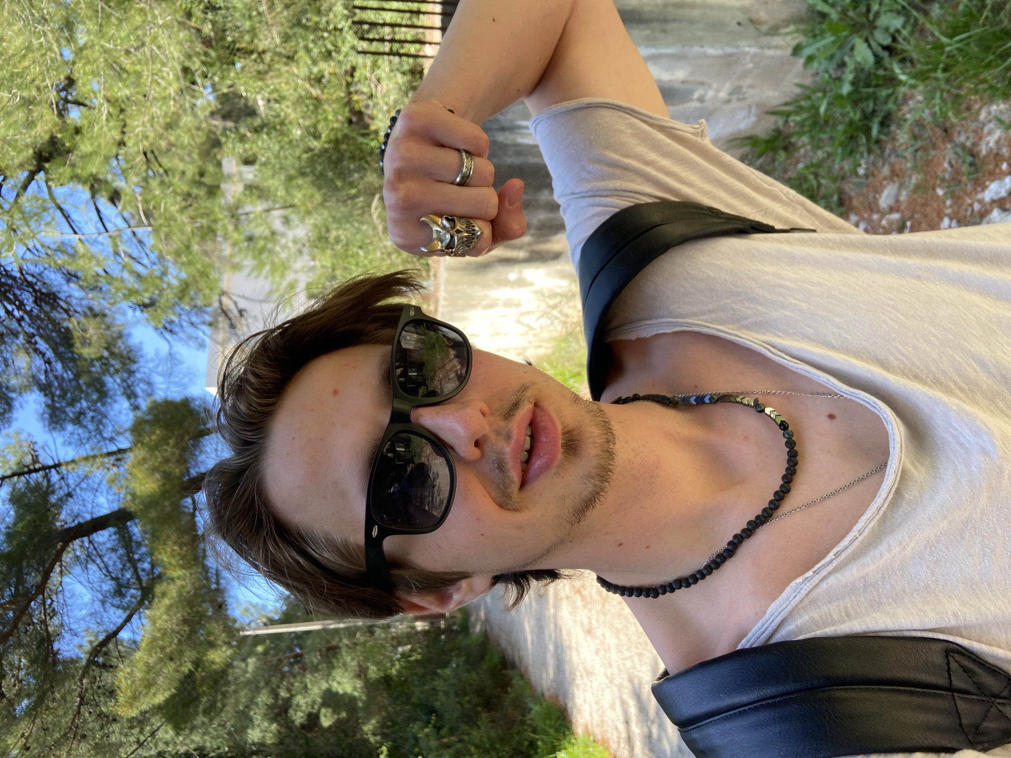 A fair-skinned man wearing sunglasses, a beige t-shirt, a black leather shoulder strap, and several rings is the primary subject. He is outdoors, likely in a park or garden. The background includes trees with green foliage and a glimpse of a light-colored building or structure. The location is Šušanj, Montenegro. The time of day appears to be daytime. The man's expression cannot be determined due to the sunglasses, but he is looking directly at the camera. The image is oriented vertically.