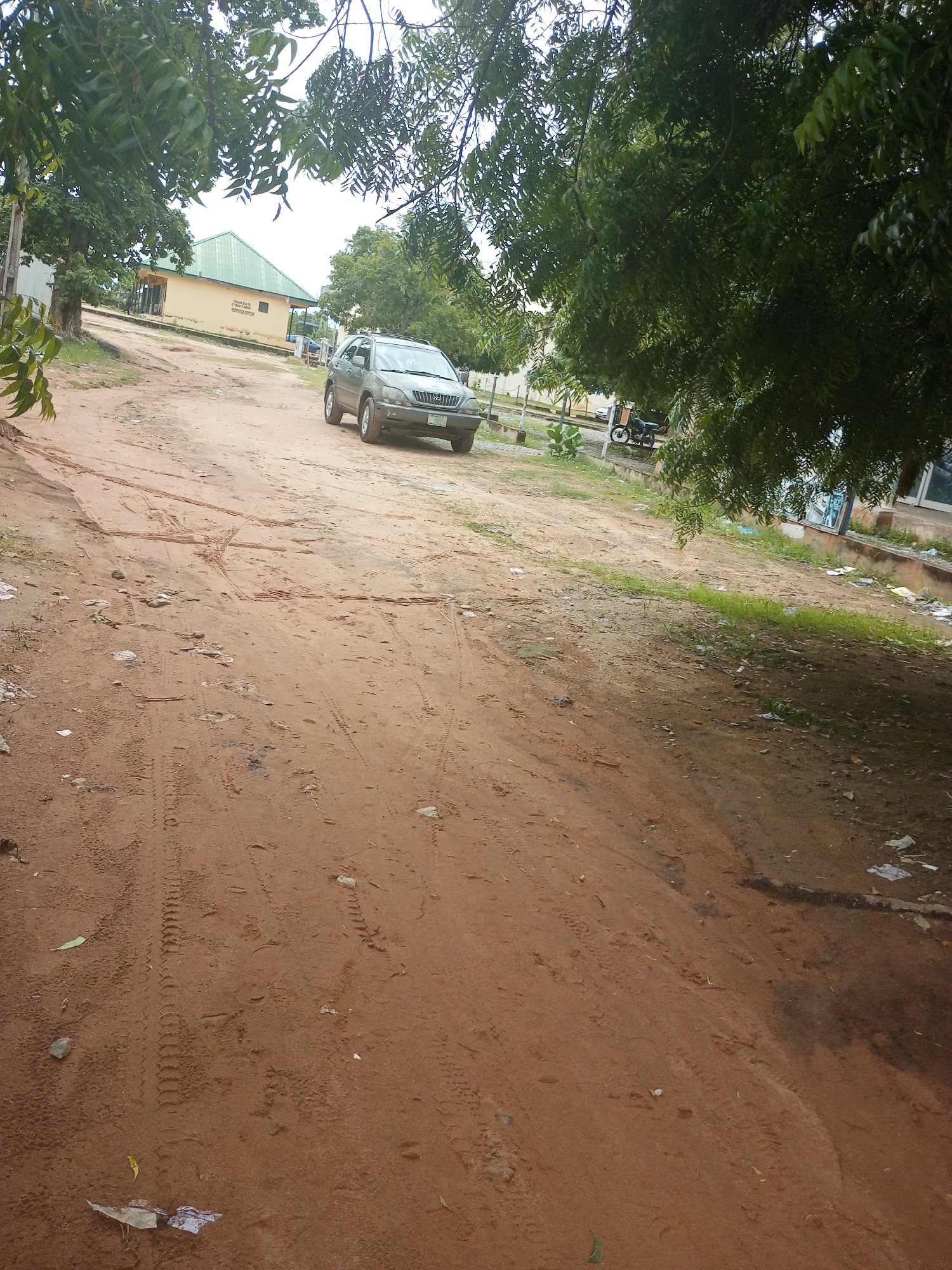 The image depicts an outdoor scene, likely on a dirt road, under an overcast sky suggesting cloudy weather or a potentially approaching rain shower. The ground is a reddish-brown dirt, marked with tire tracks from vehicles, indicating recent activity. Scattered debris, including leaves and what appears to be crumpled paper or plastic, litters the roadside.
In the middle ground, a silver SUV is parked on the left side of the road. To the left of the road, a building with a green, sloped roof and yellow walls is visible. Text on the building is partially obscured by trees but appears to include "COMPUTER CENTER". Further back, more trees and vegetation are present, along with what looks like a motorcycle parked near the building.
The foreground is dominated by the textured dirt road, with visible tire imprints and scattered debris. The overall impression is of a somewhat unkempt but functional outdoor area, possibly on the grounds of an educational institution or administrative building in Jalingo, Nigeria. The time of day is indeterminate, but the lighting suggests it is daytime. There are no people visible in the image.