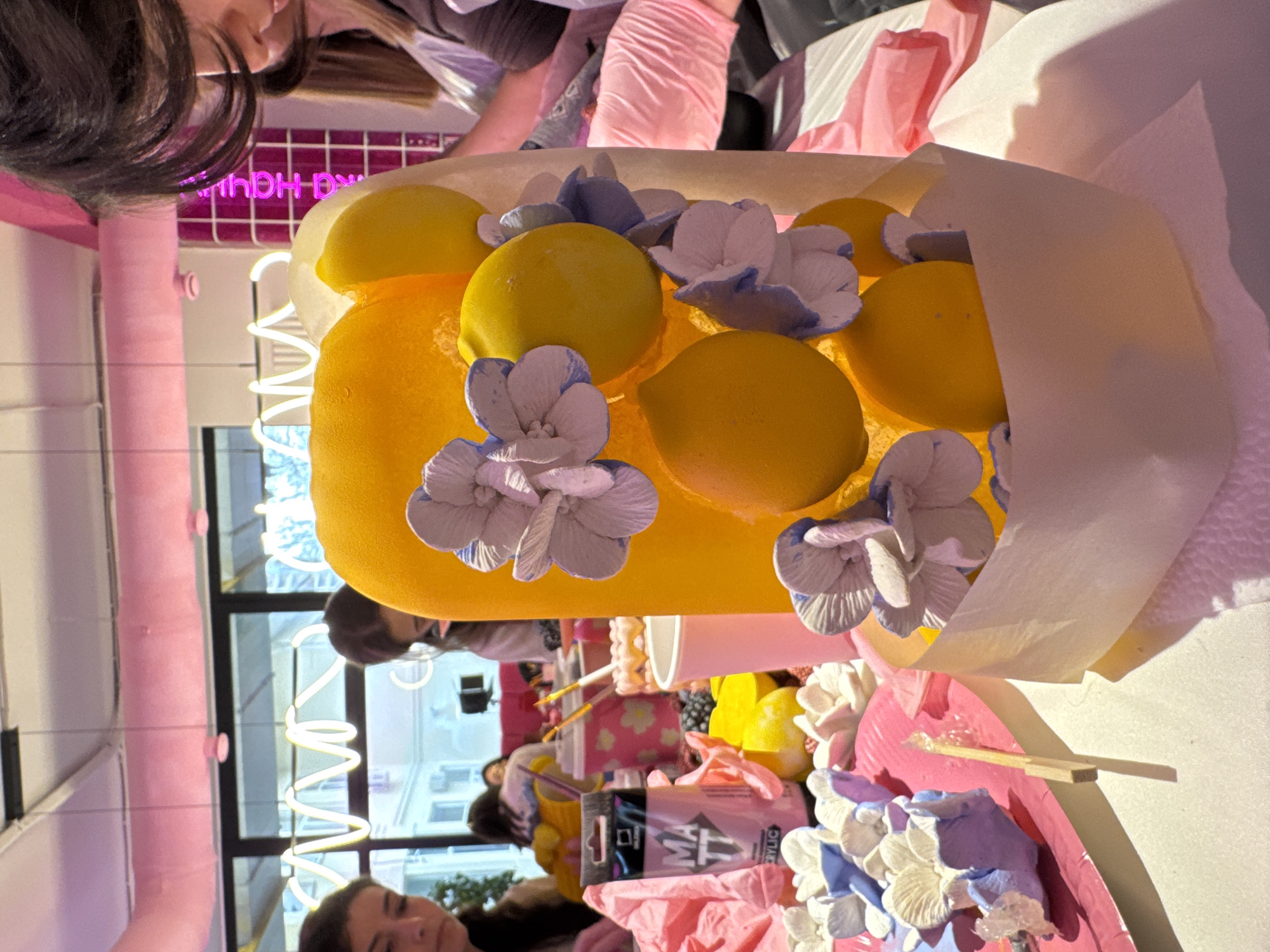 A close-up, eye-level shot captures a vibrant scene, likely indoors, with a focus on crafting and decorative elements. The primary subject is a creative arrangement featuring a collection of artificial lemons and flowers, displayed within a pale yellow wrapper. The lemons are a bright yellow, and the flowers are pastel-colored.
Surrounding the main subject, there are crafting tools and materials. In the foreground, a pink tray holds more of the artificial flowers, along with what appears to be paint tubes and other crafting supplies. People are present in the background, engaged in the crafting activity.
The environment is filled with pink accents, including a pink pillar and a pink neon sign, potentially indicating a whimsical or playful setting. Outside the window, there are buildings, suggesting an urban location. Based on the available information, it is difficult to pinpoint the exact location within Sochi, Russia, without additional data or context.