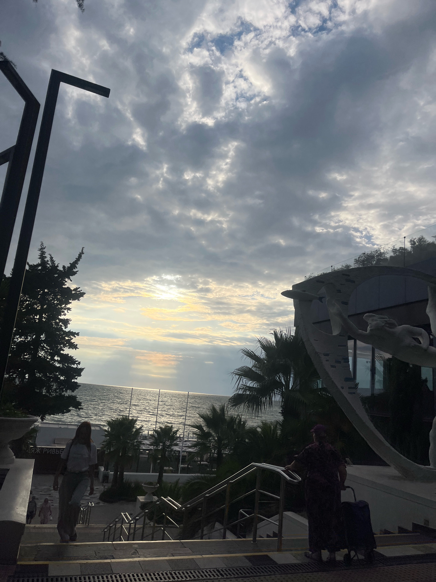 In Sochi, Russia, a cloudy sky with shafts of sunlight breaking through looms over a scene featuring a body of water, likely the Black Sea, with gentle waves. In the foreground, a woman in a light-colored t-shirt and loose pants walks down a stone staircase. Further down the stairs, another individual, dressed in dark, patterned clothing and a purple hat, pushes a dark suitcase on wheels. To the left, a silhouette of a coniferous tree stands against the sky, and to the right, a portion of a building with a curved facade and a sculpture of a human-like figure is visible. Palm trees are scattered throughout the midground, and in the distance, there are beach umbrellas and structures suggestive of a resort. The lighting suggests it is either early morning or late afternoon.