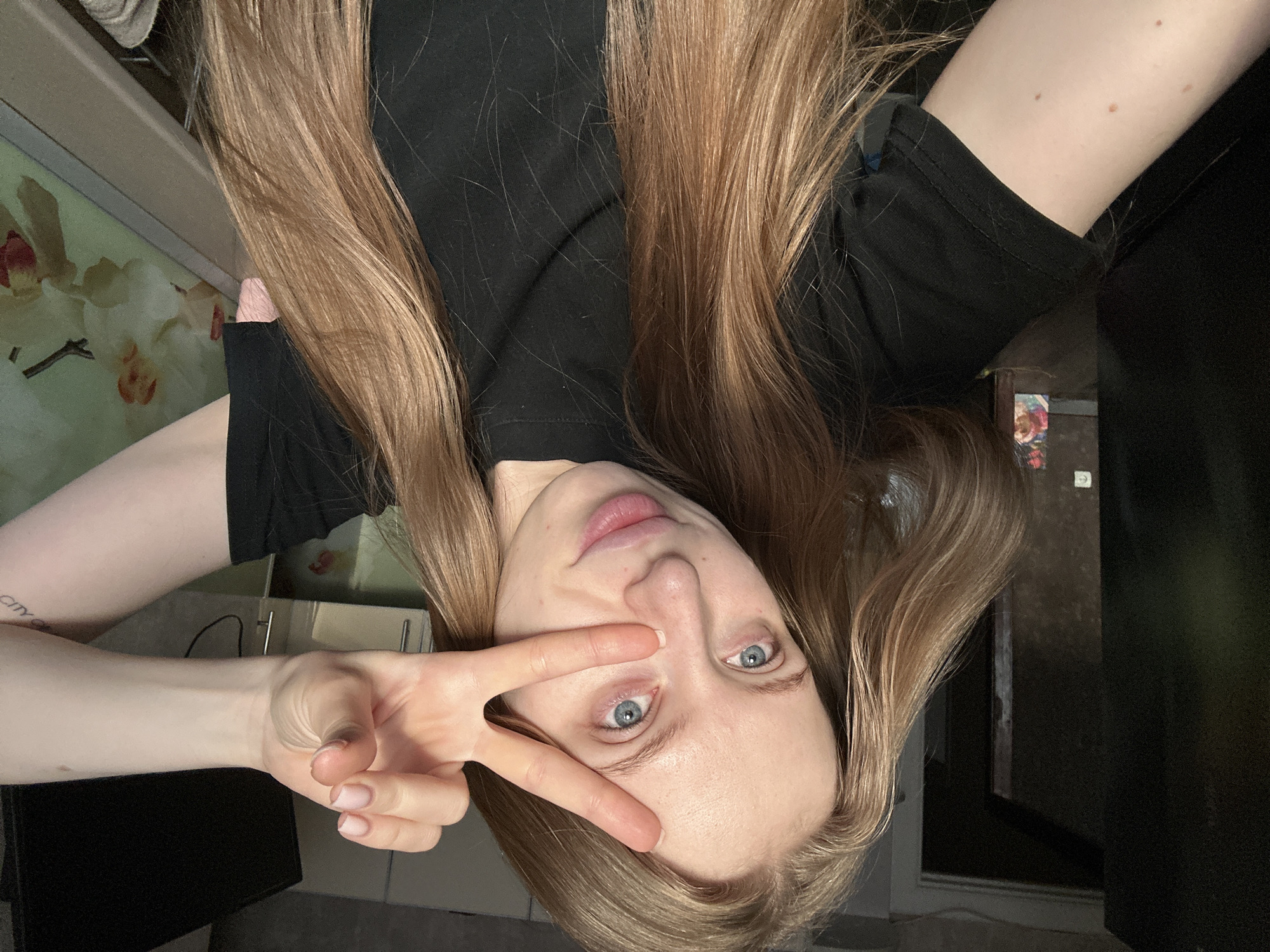 A female subject, identified by long, light brown hair and blue eyes, occupies the majority of the frame in a close-up indoor setting. She is wearing a black short-sleeved t-shirt. Her left hand, with naturally colored nails, is raised to her face, forming a "V" sign with the index and middle fingers, partially obscuring her left eye and nose. A section of her right forearm is visible on the right, bearing a faint tattoo with text, partially legible as "city." The background on the left displays a dark doorway opening into another room, where a colorful, possibly floral, artwork is visible on a distant wall. A white electrical outlet is affixed to the wall to the left of the subject. On the right side of the background, a dark rectangular object, consistent with an electronic screen, is situated above a horizontal surface. Above this surface, a decorative panel or wallpaper with a white and pink orchid pattern is present. The specific location within the city of Chaikovsky, Russia, cannot be ascertained from the visual evidence provided.