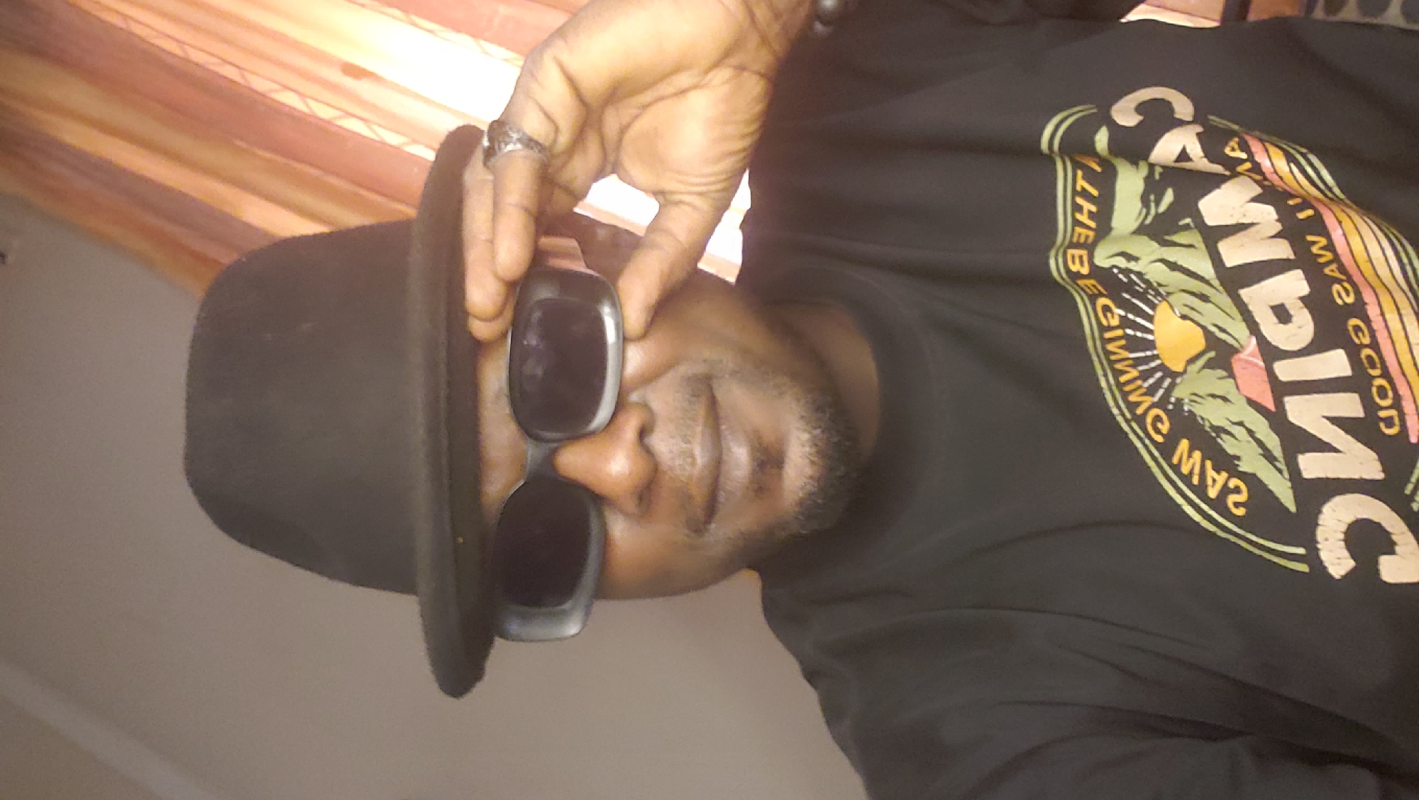 A person wearing a black fedora and sunglasses, with a ring on their finger, is wearing a black t-shirt with a circular design that reads "Saw the beginning, saw the good times, camping." The person is positioned indoors against a blurred background with orange drapery. The image was likely taken in Sabon Tasha, Nigeria.