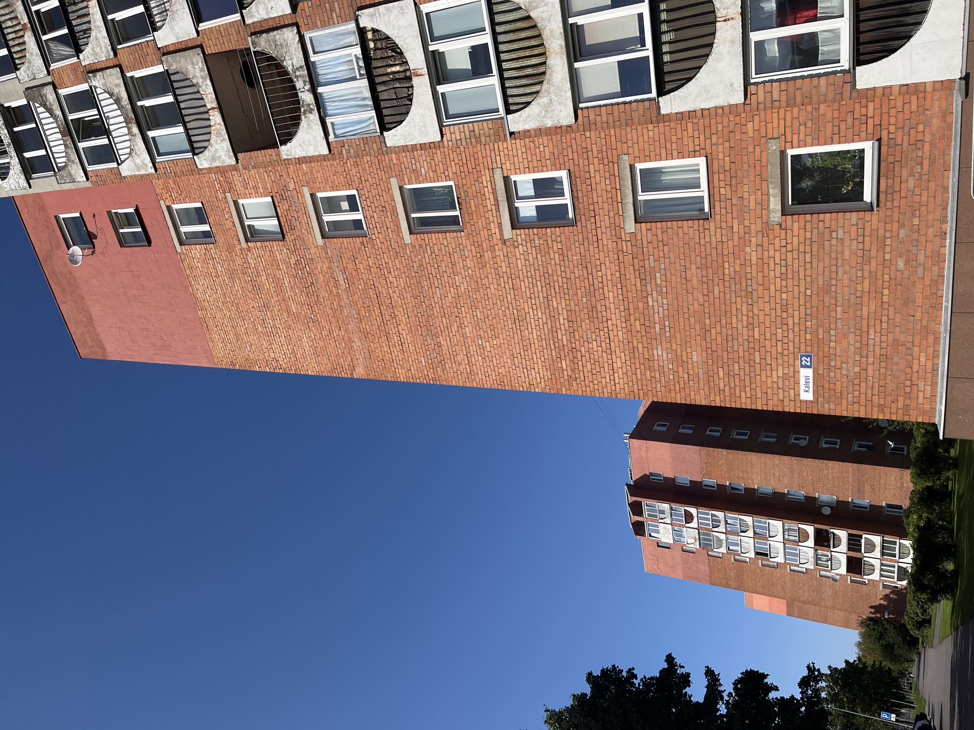 The image shows a multi-story brick apartment building with a red-painted upper section, set against a clear blue sky. The building features numerous windows and balconies with arched concrete fronts. A street sign reading "Kalevi 22" is visible on the brick facade. To the left of the main building, another similar apartment building is partially visible, also constructed with red brick and having a similar architectural style. The foreground shows a paved walkway and a grassy area with some trees and bushes. The scene appears to be a residential area in a city, under bright daylight.