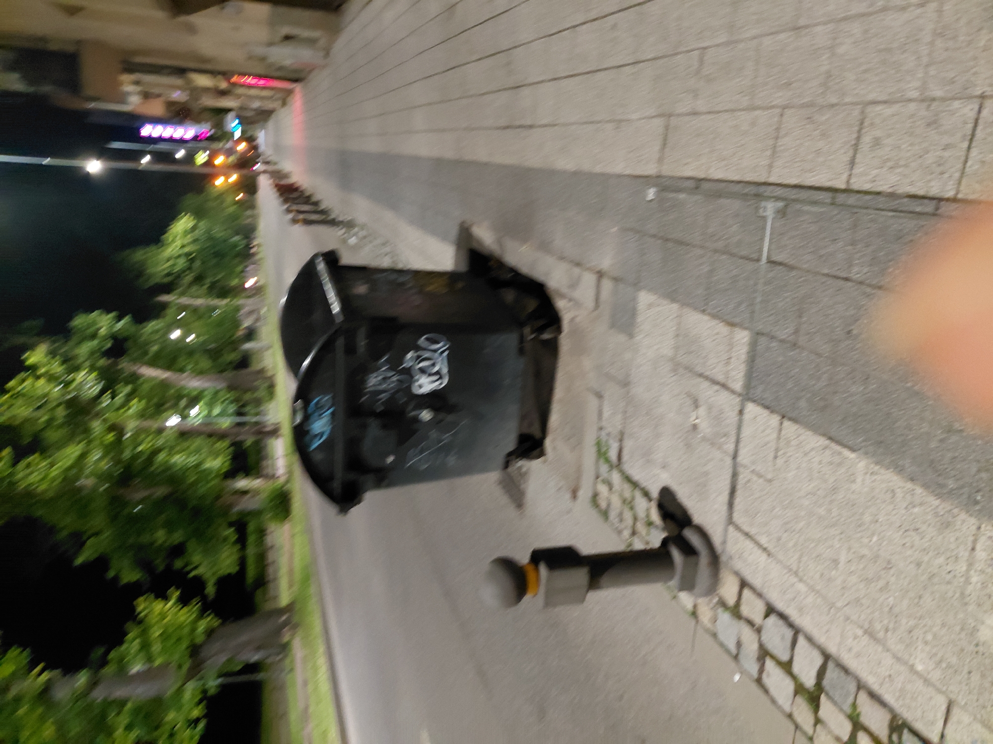 This nighttime image depicts a street scene in the city of Plovdiv, Bulgaria. The perspective is low, looking down a street with a prominent sidewalk on the right. In the immediate foreground, a blurry peach-toned object, possibly a finger, partially obstructs the lower right corner. The sidewalk is paved with light grey rectangular slabs, bordered by a darker grey strip and a section of cobblestones near the curb. A dark grey metal bollard with a spherical top and a single orange reflective band is anchored in the cobblestones on the left side of the sidewalk.
In the midground, two large, dark grey industrial trash bins with curved tops are situated on the sidewalk, adjacent to the curb. Both bins display white, abstract graffiti markings, including a stylized "OZO" or "020" on one. A line of simpler, dark metal bollards extends further down the sidewalk, separating it from the asphalt road on the left. The road, dark grey in color, runs parallel to the sidewalk.
The left side of the background features a row of large trees with green foliage, bordering the road. Several streetlights emit bright white-yellow light among the trees. The sky is dark, consistent with a nighttime setting. On the far right, buildings with light-colored facades line the street, displaying various colorful neon signs, including a prominent vertical stack of pink/purple rectangular lights, indicating commercial activity further down. Another bright street light illuminates the distant sidewalk and road on the right. No people or specific actions are visible in the scene.
