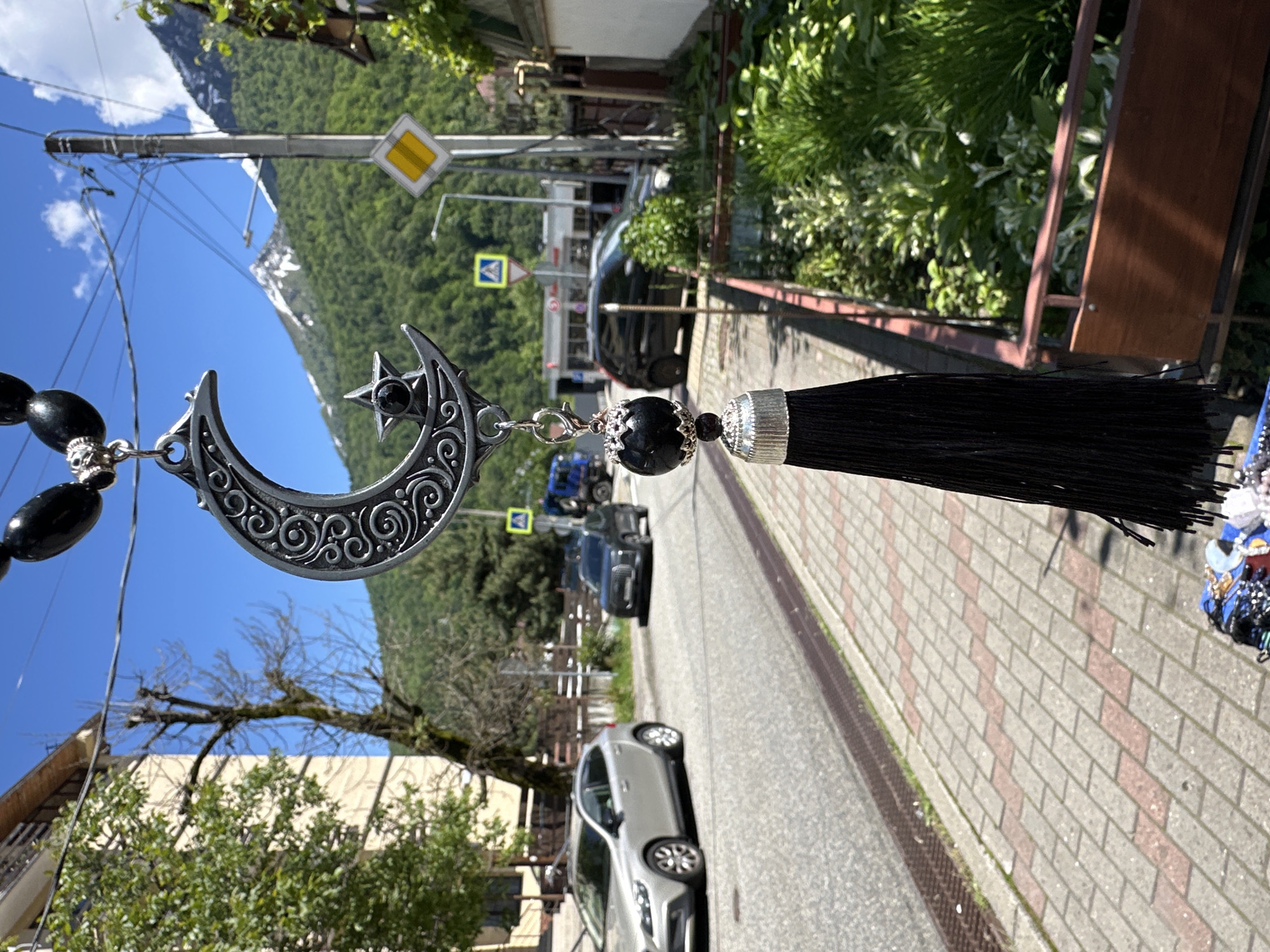 The image captures a detailed silver-colored crescent moon and star charm with black beads and a black tassel, positioned prominently in the foreground. The charm features intricate patterns and a black gemstone set within the star. It appears to be hanging, possibly from a car's rearview mirror or a display.
The background reveals a sunny, mountainous landscape in Krasnaya Polyana, Russia. Lush green, forested mountains rise majestically, with glimpses of snow-capped peaks in the distance under a clear blue sky.
A paved road with cars (including a silver hatchback and a dark SUV) runs through the scene, bordered by a patterned sidewalk made of light and reddish-brown bricks. Utility infrastructure, such as a tall pole with power lines and a street lamp, is visible. Road signs include a yellow diamond (indicating a priority road) and a blue square sign with a white arrow, likely for direction or mandatory turn. Various trees and greenery line the street, alongside buildings with balconies. The bright light and strong shadows suggest it is midday or early afternoon.