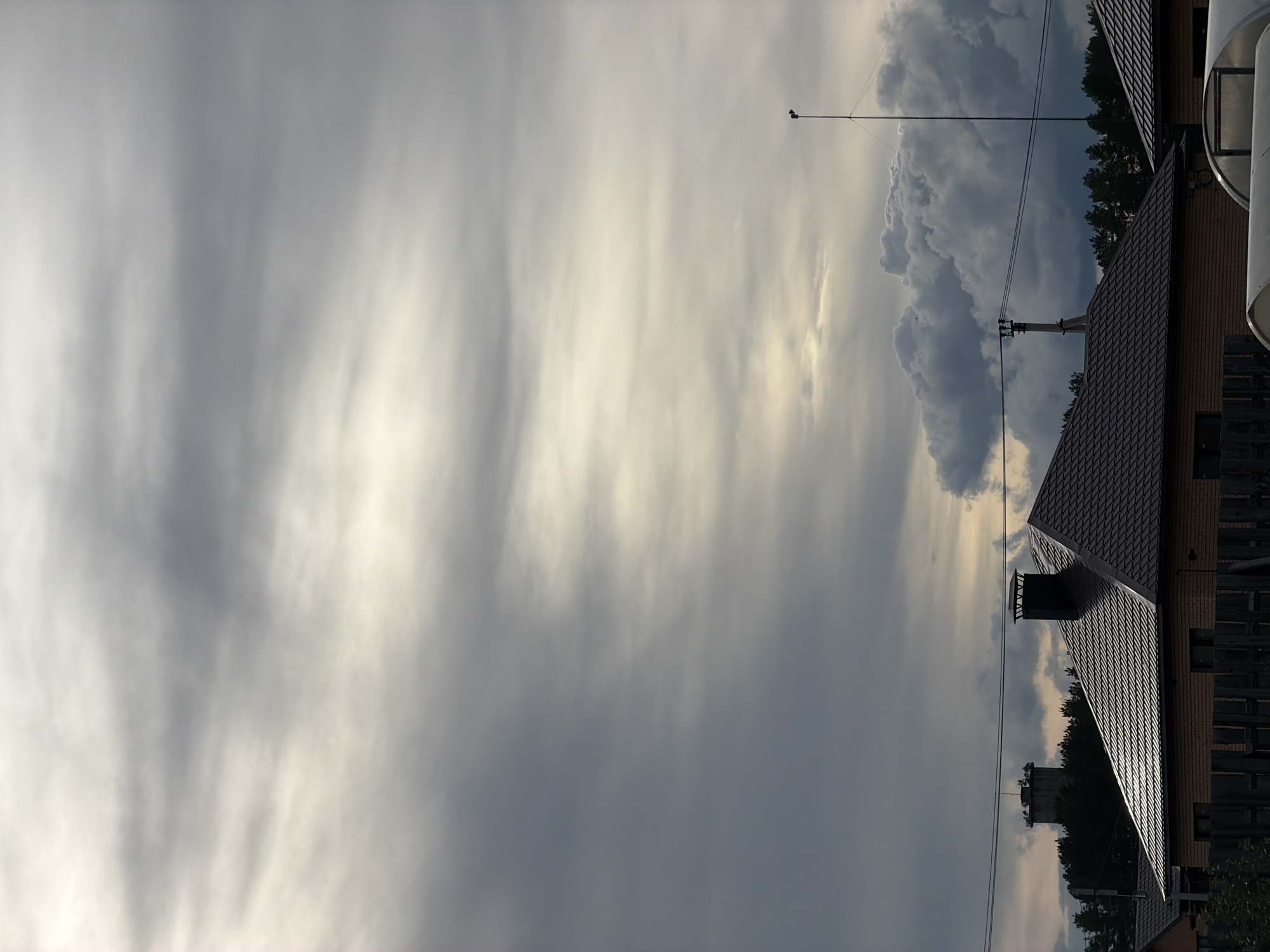 The image depicts a cloudy sky above rooftops and trees in Chaikovsky, Russia. The sky is predominantly gray and white with striated cloud formations, hinting at possible weather disturbances. A transmission tower is visible in the upper portion of the image. Power lines cross the frame. The roof of a brick building with dark brown tiles is prominently displayed. Trees are visible in the background, suggesting a suburban setting. A portion of a white cylindrical structure is observed on the building to the right.