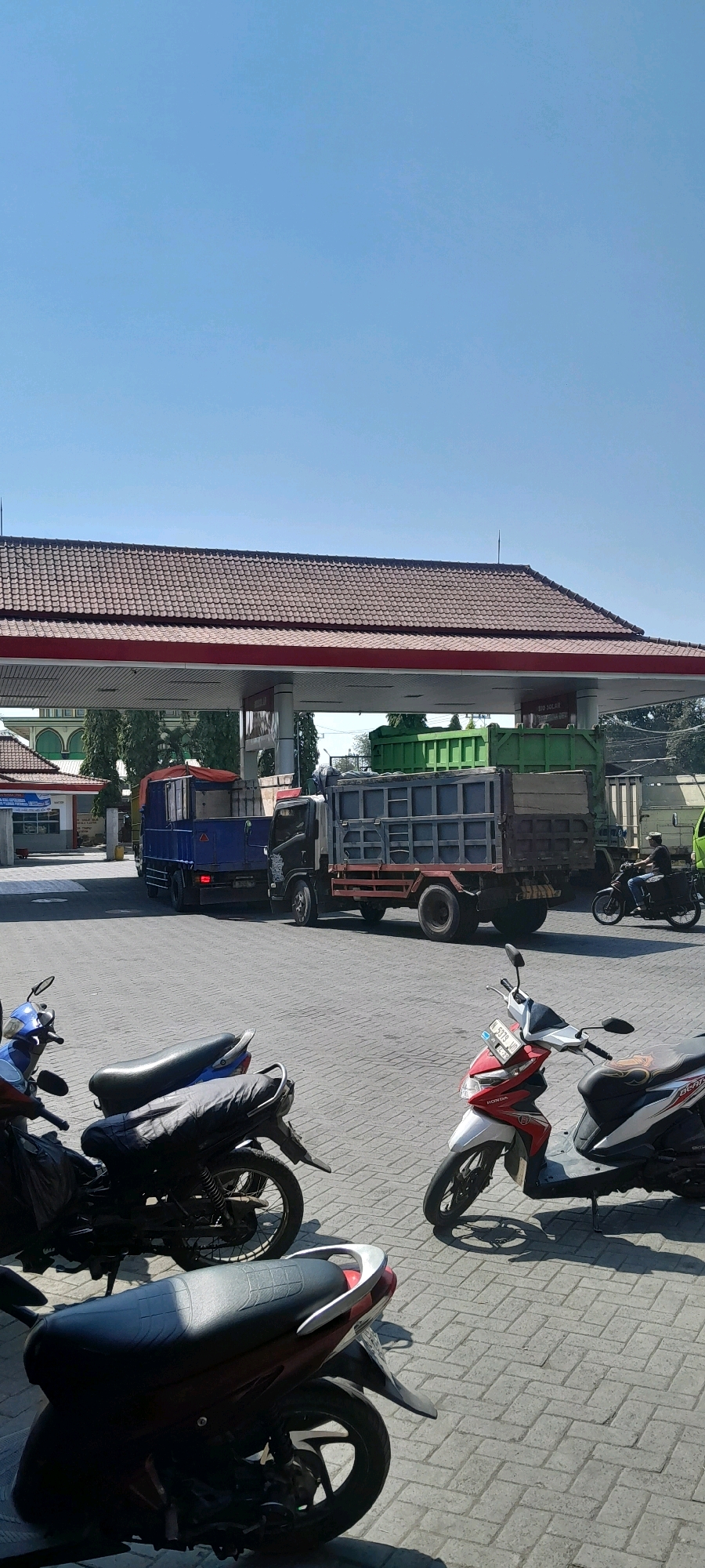 The image depicts a sunny outdoor scene at a gas station in Semare, Indonesia. In the foreground, several motorcycles are parked on a brick-paved surface. Two are a deep red and black, one is mostly black, and another is blue. A red and white scooter is also present. In the mid-ground, two large trucks are visible. One is a blue truck with a red tarp covering its load. Next to it is a grey flatbed truck with a metal cargo area, and behind that, a green truck bed. A person is seen riding a dark motorcycle in the background, moving from right to left. The gas station features a red-roofed canopy supported by white pillars, with signage visible on one of the pillars. In the background, trees and a building with a green roof are present. The sky is a clear, bright blue.