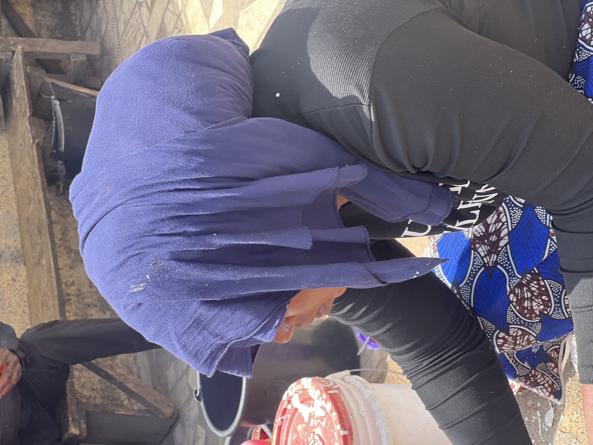 An overhead shot captures a person, likely female, in a sunlit outdoor or semi-outdoor setting in Jos, Nigeria, bending forward as if engaged in work. The central subject wears a deep blue head covering, obscuring most of their face, though a glimpse of their nose with a small blue stud is visible. They are dressed in black ribbed trousers or leggings, layered with a vibrant wrapper or skirt featuring a blue, brown, and white patterned design.
The person is positioned near several buckets, one prominent white and red bucket splashed with a light-colored substance, possibly paint or plaster, suggesting an activity involving liquid or semi-liquid materials. A dark-colored bucket is also seen nearby. In the background, to the left, wooden steps or a wooden structure are visible, and the ground appears to be paved with light-colored tiles or concrete. The leg of another person, wearing black trousers, is partially visible in the lower-left corner of the frame. The scene is brightly lit, indicating daytime conditions. No discernible text is visible within the image.