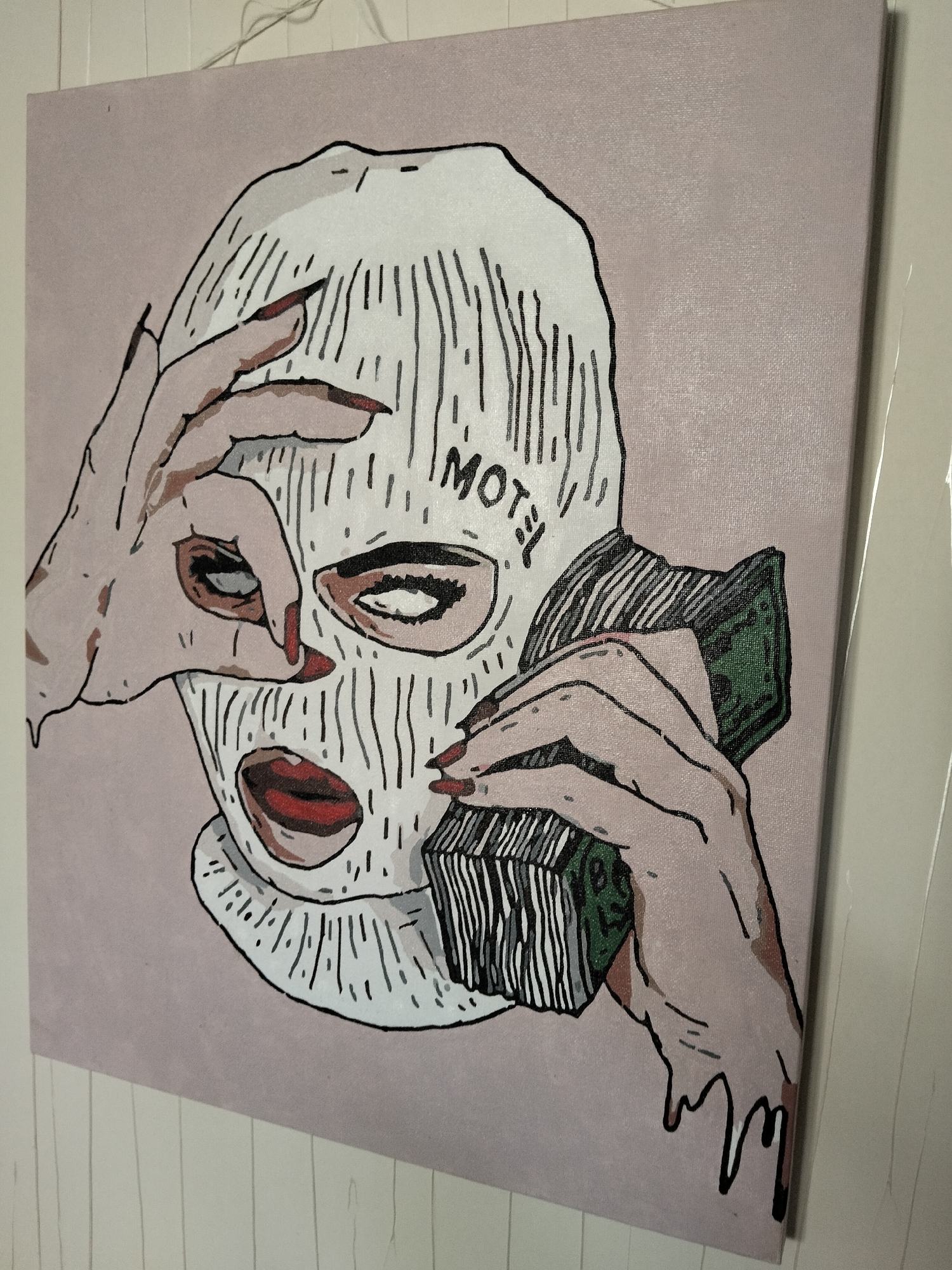This is a painting on canvas of a person wearing a white ski mask and holding a wad of cash to their ear as if on a phone. The mask has black lines drawn to simulate knitting and two eyeholes. The word "MOTEL" is written in black lettering on the mask above the right eyehole. The person has red lipstick and manicured red fingernails, visible on the hands holding the money and peering through one eyehole. The background is a solid, muted pink. The painting is hung with a thin string.