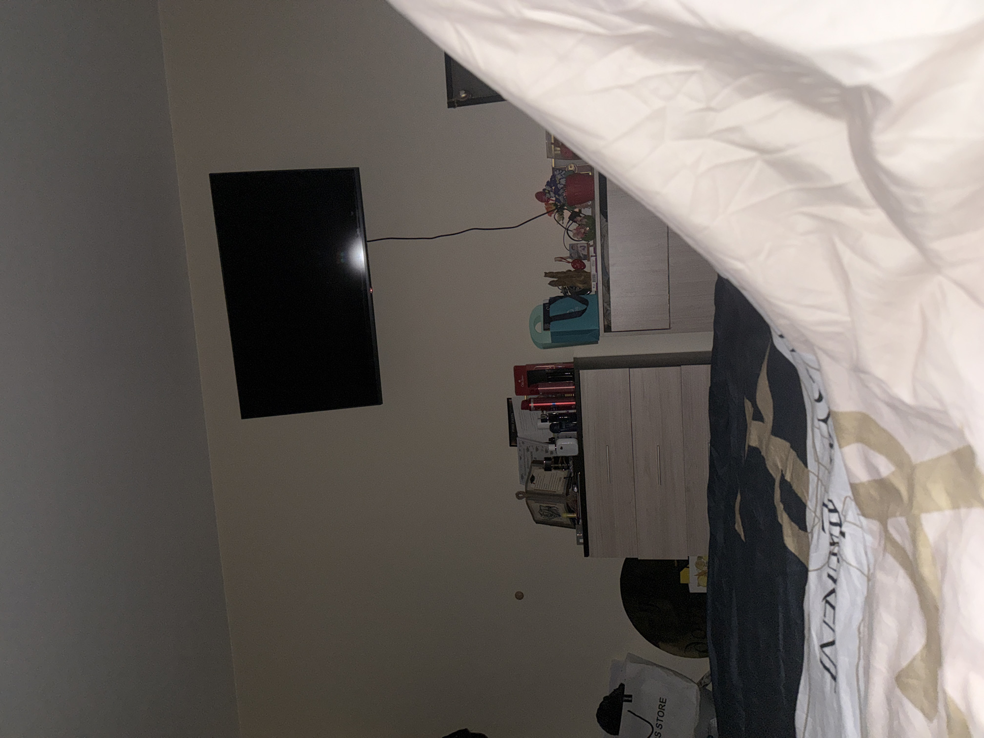 The image shows a room with a television mounted on the wall. Next to the TV, there is a cabinet or shelf with various items. A bed or bedding is partially visible in the foreground.
