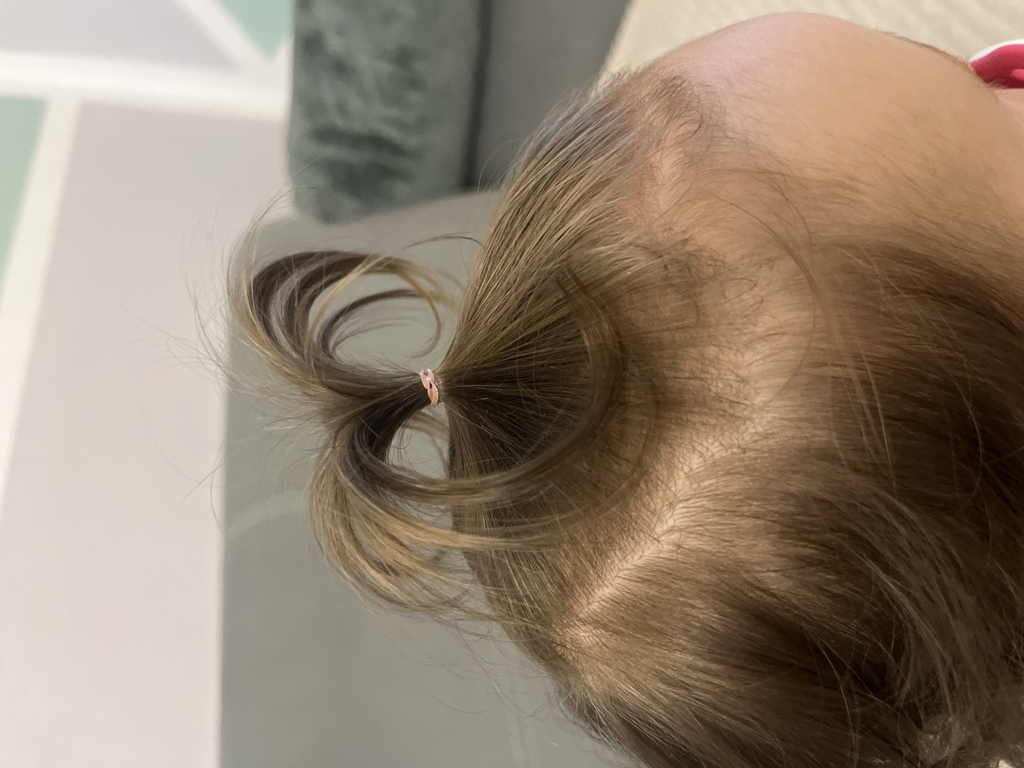 A close-up view focuses on the top of a toddler's head with a small ponytail secured with a pink hair tie. The hair is light brown with some lighter strands. The skin on the scalp is visible. The background includes a wall with a geometric pattern in shades of green and white. The location is Unknown, Unknown.
