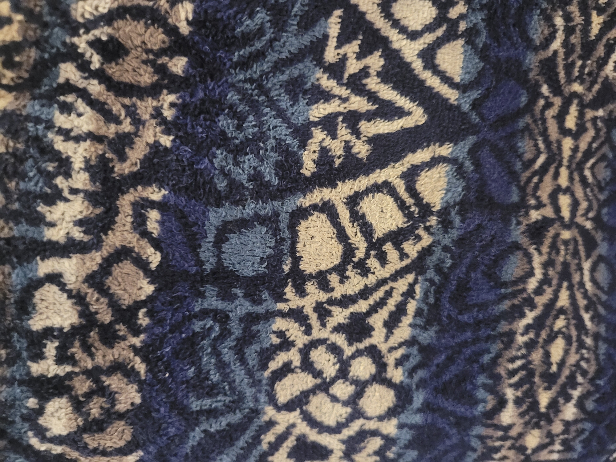 The image shows a close-up view of a textured fabric with a repeating abstract pattern in shades of dark blue, light blue, beige, and brown. The pattern consists of irregular shapes and lines that resemble organic forms or stylized animal prints. The fabric appears to be thick and possibly plush, such as a carpet or upholstery. There is no indication of any specific location in Gävle, Sweden, or any people, actions, or interactions.