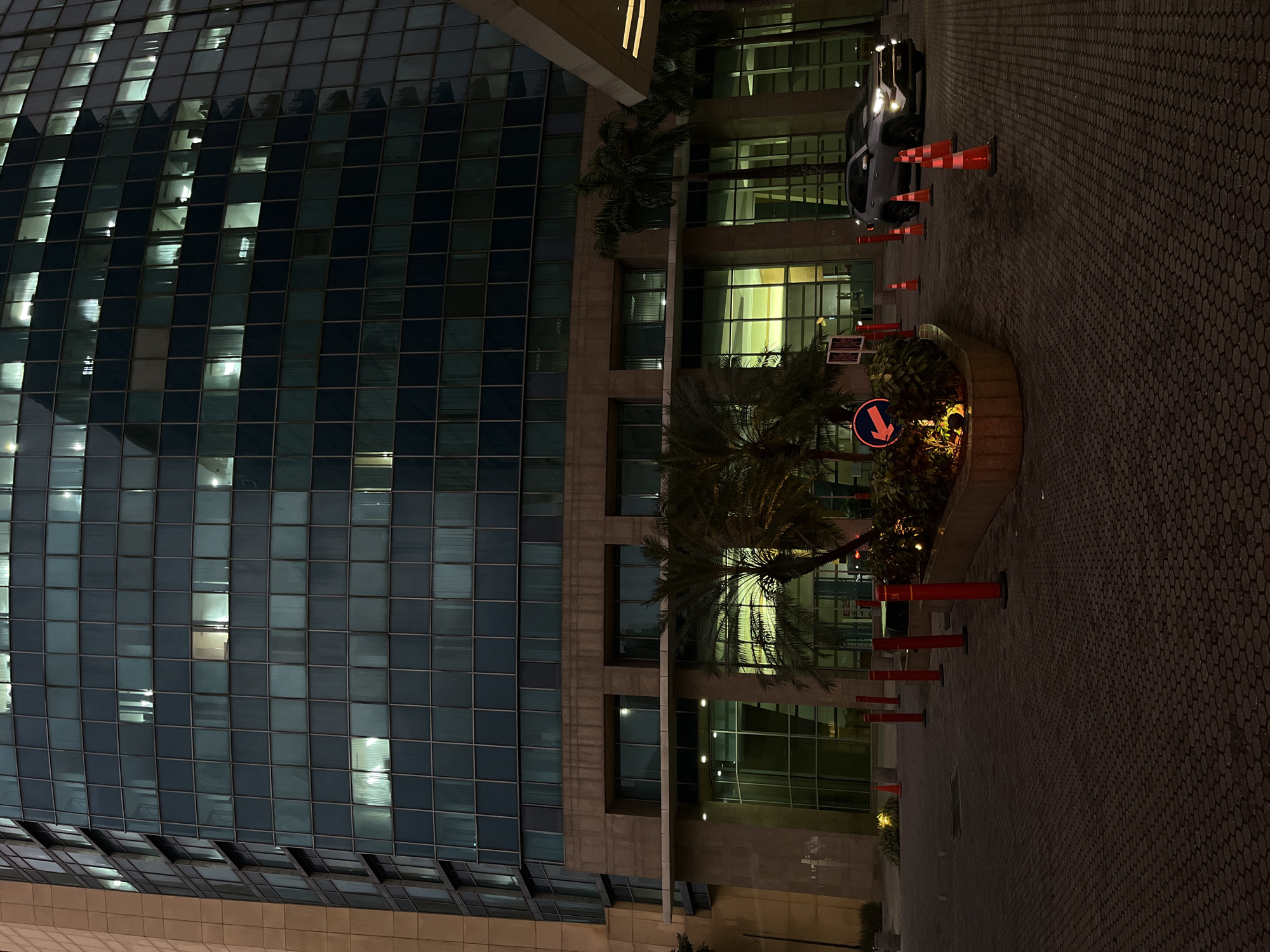 The photograph shows the exterior of a building, possibly an office or commercial structure, at night. The building has a modern design with a facade dominated by a grid of windows, some illuminated from within, suggesting activity inside. To the right of the building, there's a street or driveway paved with hexagonal tiles, with several orange traffic cones and red bollards lined along the edge. In the center, a circular flower bed is visible with a sign and some vegetation, lit from below. A car is parked in the background, facing the building. The overall impression is of a quiet urban scene at night. The location is Unknown, Unknown.