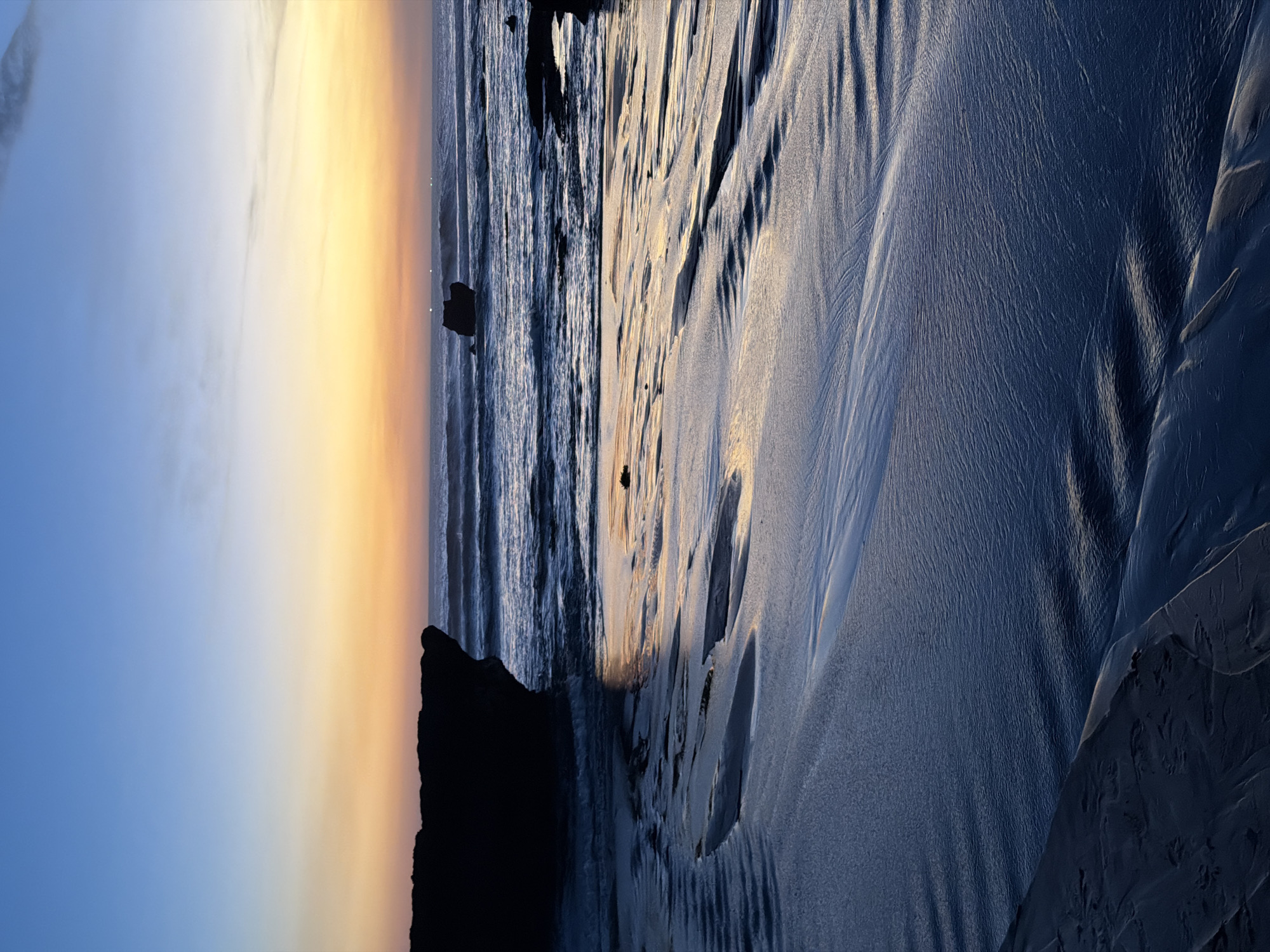The image depicts a beach scene at either sunrise or sunset, with the sky transitioning from a deep blue on the left to a warm orange and yellow gradient towards the right. The sea is visible on the right side of the frame, with small waves lapping at the shore. The foreground is dominated by wet sand, reflecting the colors of the sky and showing intricate patterns left by the receding tide. Several dark, rocky outcrops are present along the waterline. The overall mood is serene and tranquil, enhanced by the soft lighting and natural textures of the beach. There are no visible people or text in the image. The location is North Kuta, Indonesia.