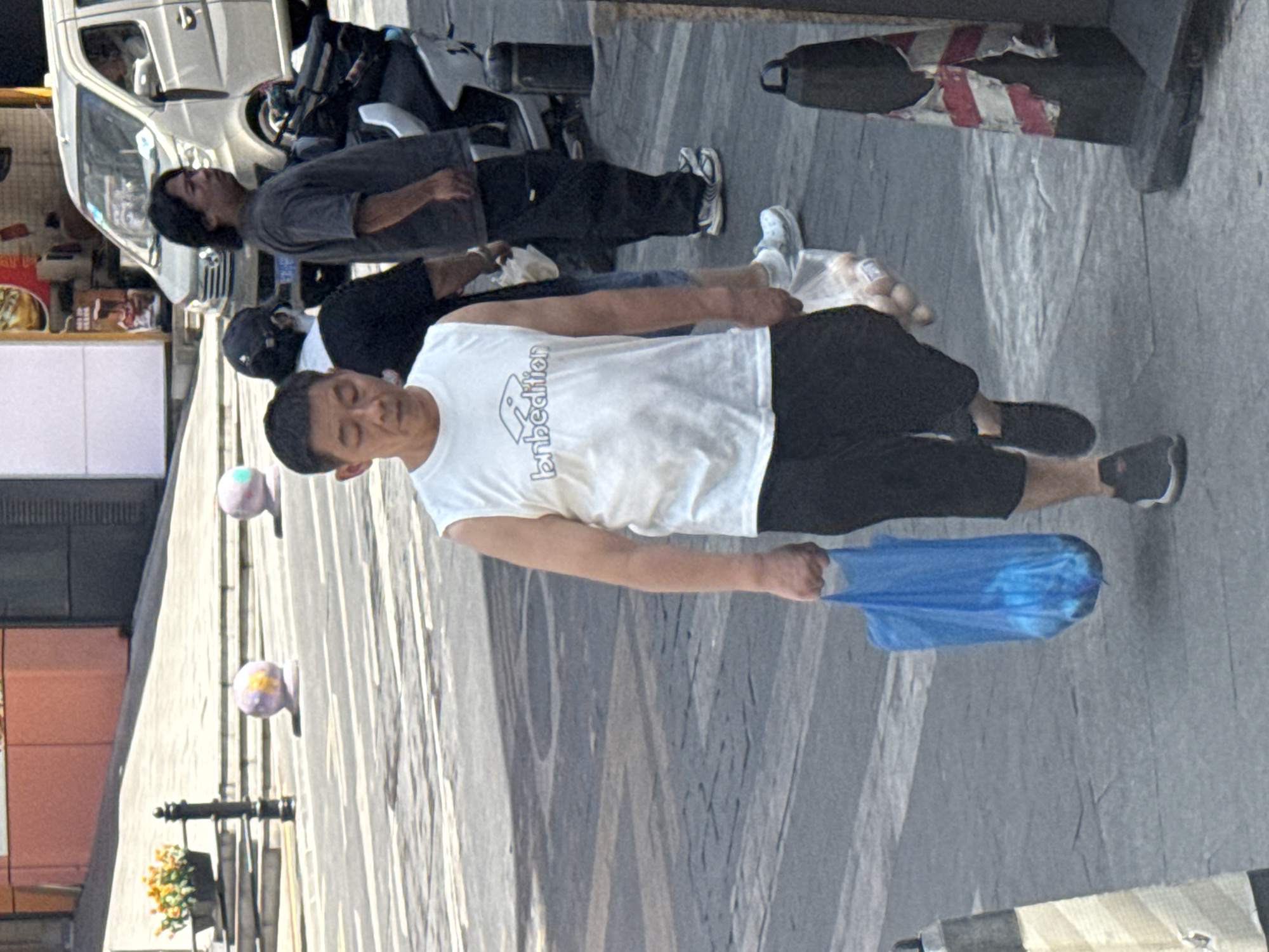 A middle-aged East Asian man walks across a paved street in Yuzhong District, China, carrying a blue plastic bag in his left hand and a clear plastic bag containing round objects in his right hand. He is wearing a white sleeveless t-shirt with a logo that appears to say "bnbedition" and dark shorts. His feet are clad in black and white athletic shoes. Behind him, a man with dark hair and a dark t-shirt stands near a parked silver minivan. Another person wearing a black baseball cap and a white collared shirt is partially visible between them. The street is marked with white lines and the background shows a building with orange and brown facades, a lamppost, and decorative spherical objects on the sidewalk. Traffic cones are also present on the side of the street. The overall scene depicts a street-level view of urban activity under daylight.