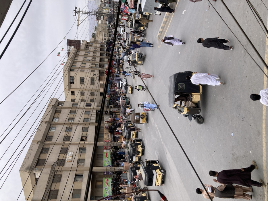 The image depicts a busy street intersection in Quetta City Tehsil, Pakistan, under an overcast sky. Multiple multi-story, light-colored concrete buildings line the street, their ground floors featuring commercial establishments with awnings and signs displaying Urdu script. Overhead, a dense network of black utility lines crisscrosses the frame. The paved road is actively used by numerous vehicles, predominantly black and yellow auto-rickshaws, along with motorcycles and a few cars. A gray truck is visible on the right. Numerous individuals, primarily adult males in traditional attire, are observed walking on the street, some operating vehicles. Three small flags, colored green and white, are visible atop a building in the mid-ground. Distant mountains are present on the horizon.