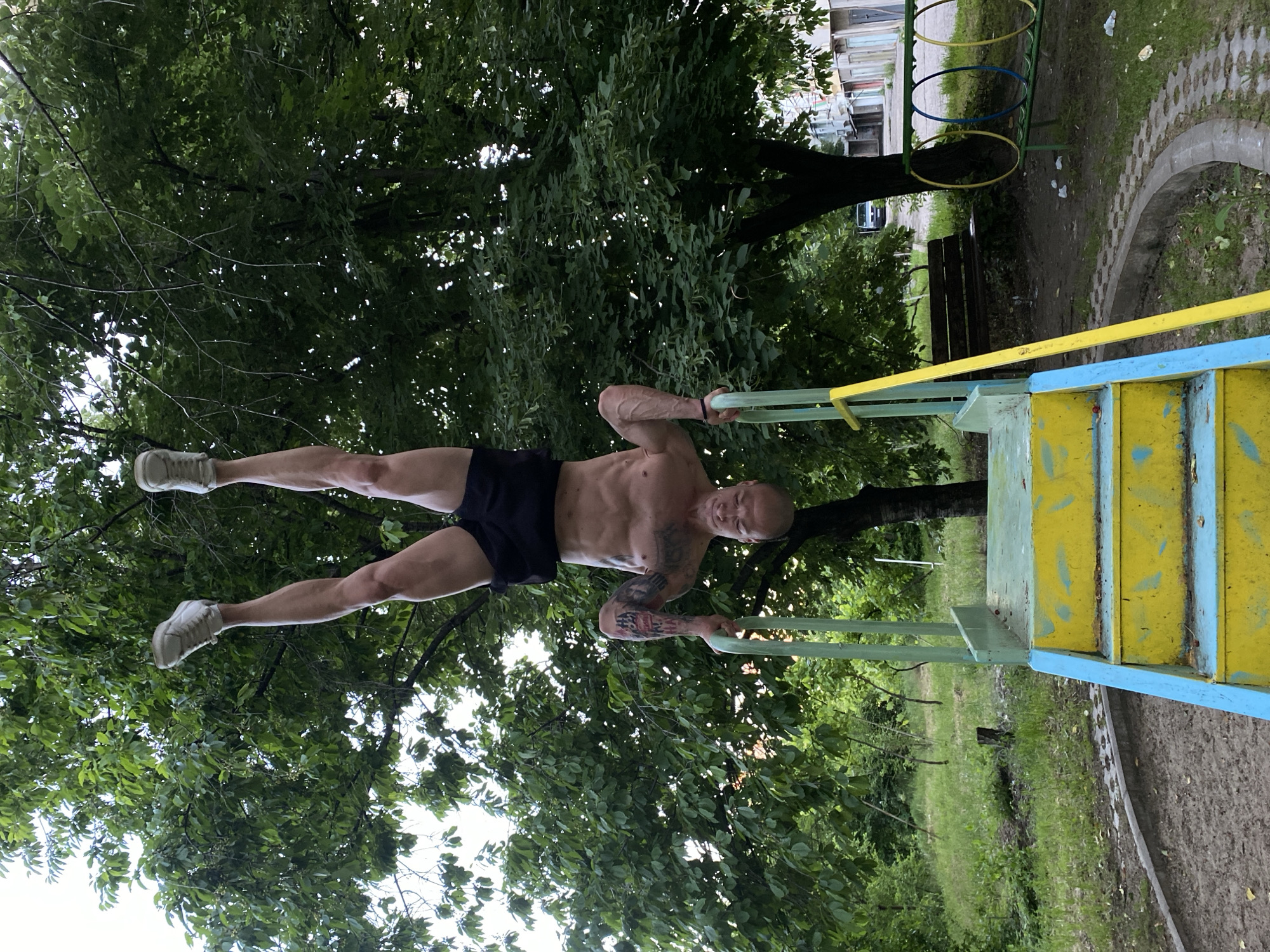 A fit, muscular, bald man with tattoos is performing a calisthenics exercise on a painted metal playground structure in a park. He is positioned horizontally, holding onto parallel bars, with his body and legs straight and parallel to the ground. He wears dark shorts and white sneakers. Green trees and foliage surround the playground structure. The park appears to be in Pleven, Bulgaria, as indicated by the architecture of buildings in the background. Other playground equipment, including colored rings hanging from a structure, are visible nearby.