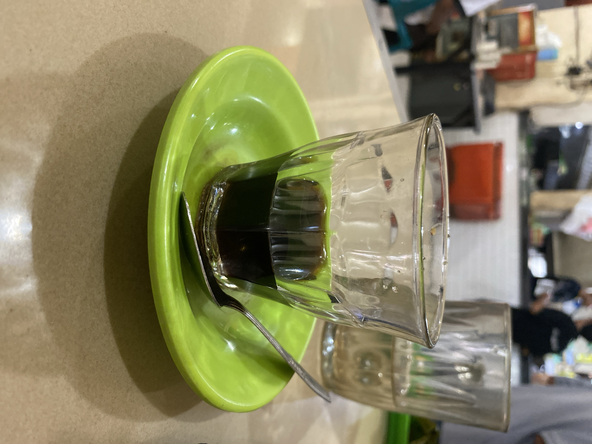 A close-up, eye-level shot captures a small glass filled with dark liquid, likely coffee, sitting on a bright green saucer. A silver spoon rests on the saucer, positioned partially under the glass. In the background, a second, larger glass of clear liquid is visible, along with blurred figures of people and an indistinct environment that suggests a casual eatery or cafe. The table surface is a neutral beige. The setting appears to be a local establishment in Lhokseumawe, Indonesia, possibly a warung kopi, a popular type of Indonesian coffee shop.