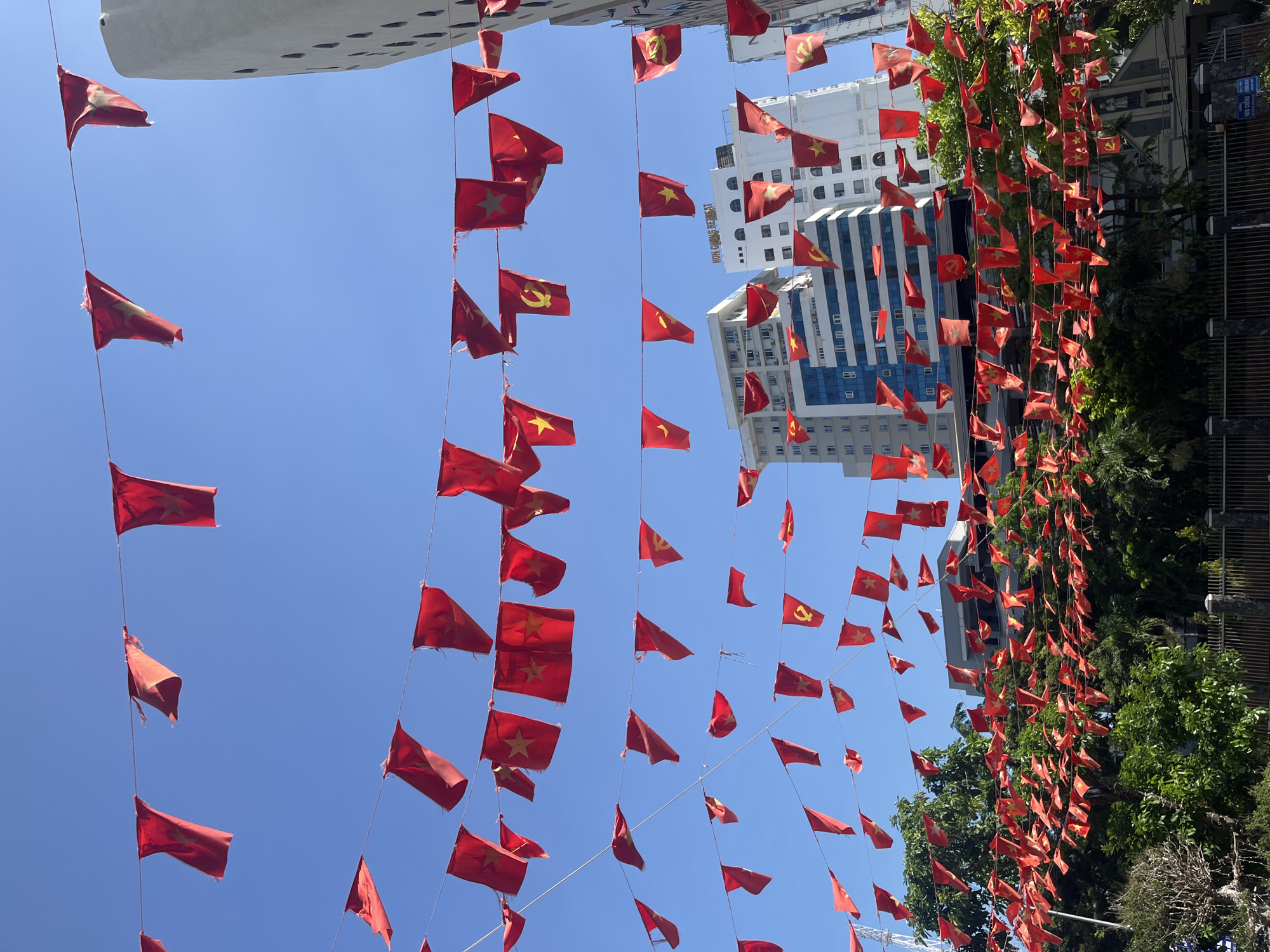The image depicts a daytime outdoor scene in Bac Nha Trang Ward, Vietnam, under a clear blue sky. Numerous small, red, triangular pennants are strung across multiple parallel wires spanning the frame. The majority of these pennants feature a yellow five-pointed star, representing the national flag of Vietnam. Several pennants also display a yellow hammer and sickle emblem. The flags appear somewhat worn. In the background, multiple multi-story buildings are visible, including a prominent white structure with "HON CHONG RETREAT" inscribed on its upper facade. Other buildings display a mix of modern architecture with glass windows and some with distinctive circular or arched window patterns. The lower portion of the image is dominated by lush green foliage, consisting of various trees and bushes. A dark metal fence with concrete posts runs along the bottom edge, separating the vegetation from an unseen ground level. A blue and white street sign with Vietnamese text, partially visible in the bottom right, reads "HOA CHUNG" on one line. No people or vehicles are present in the scene.