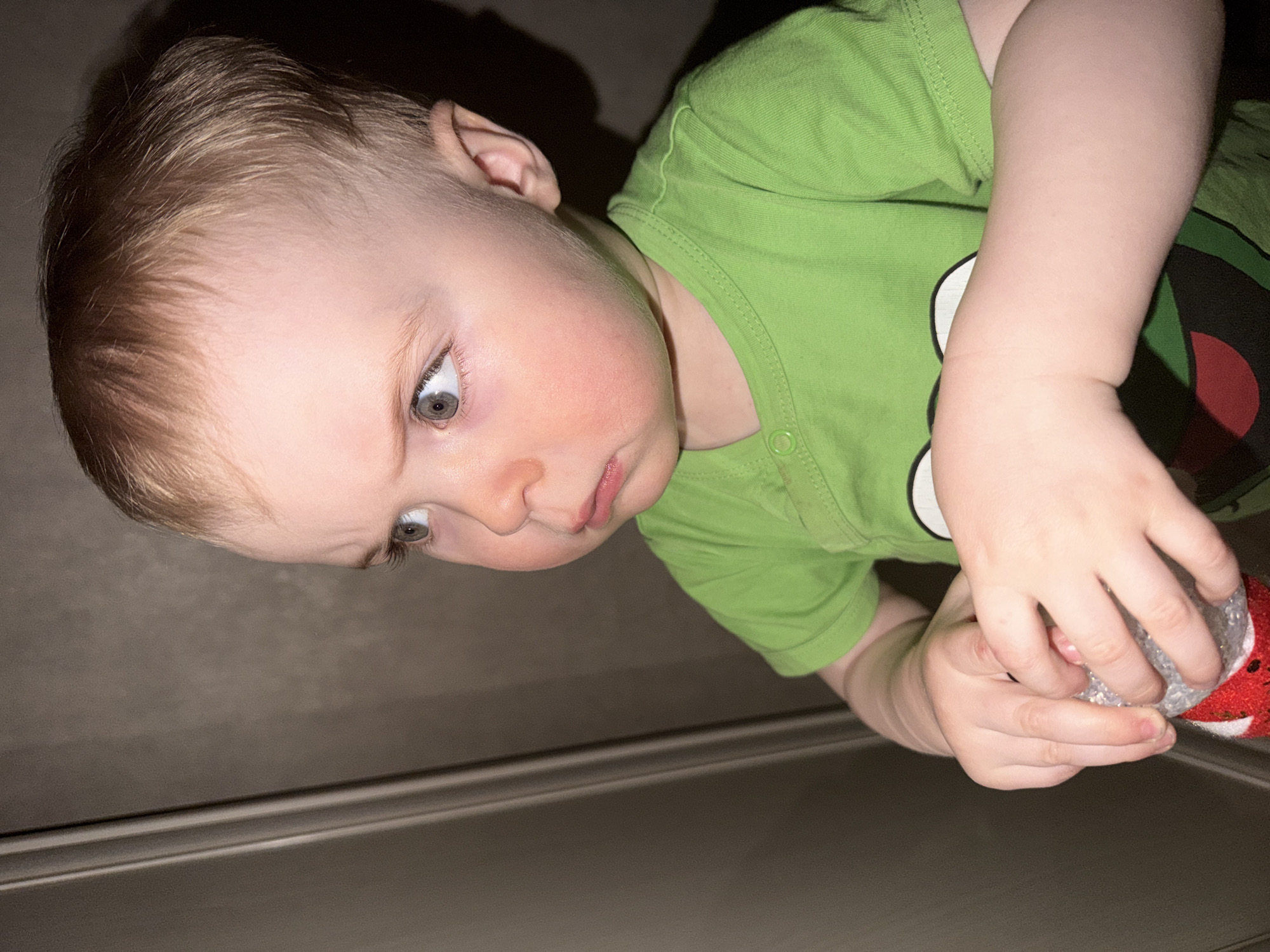 A child wearing a green shirt with a circular graphic is holding a multi-colored object with their hands. The child is positioned near a metallic surface with distinct horizontal lines. The location is unknown, and there is no indication of it being in the city of Votkinsk, Russia.
