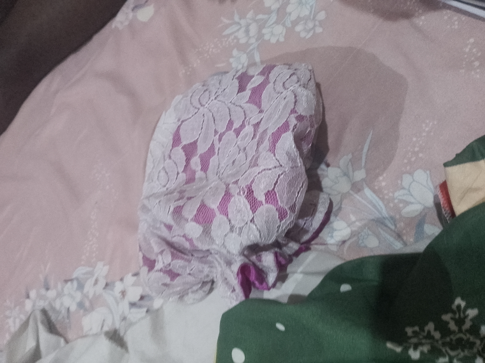 The image depicts a close-up, overhead view of intimate apparel, specifically a pair of lace panties with a pink lining, resting on a pink floral-patterned fabric, possibly a bedspread. The panties are bunched up and positioned slightly off-center to the left of the frame. The fabric beneath them has a soft pink base with a delicate white floral pattern. To the right of the panties, a portion of a dark green garment is visible, also with a pattern of white shapes. The lighting is subdued, creating some shadows on the fabric. The overall impression is of a domestic, personal setting. No specific location in the city of Unknown or any people are discernible.