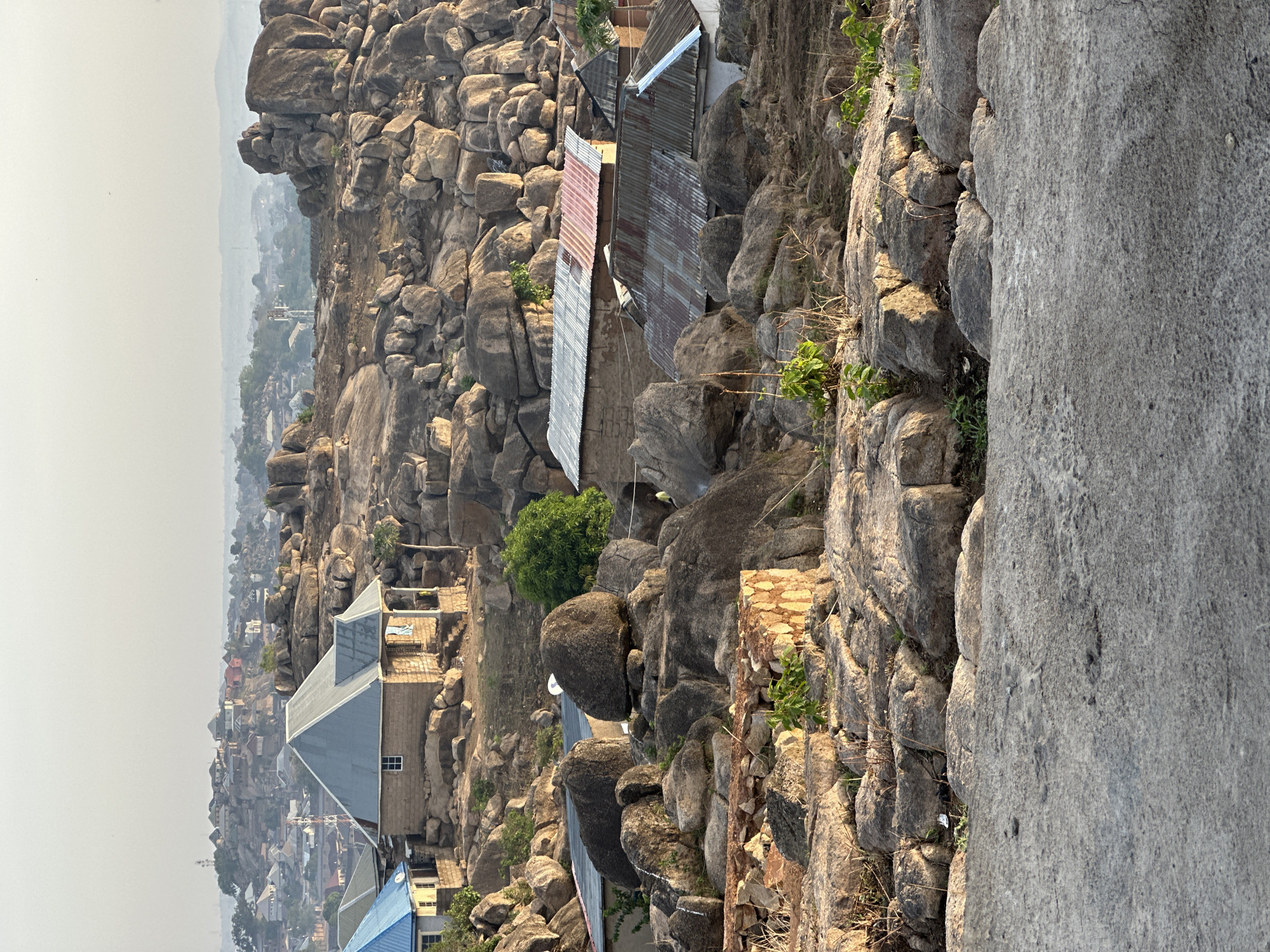 The image displays a rugged, rocky landscape in Jos, Nigeria, featuring human settlements integrated within natural boulder formations, with a hazy cityscape in the distance. The scene is dominated by massive, weathered grey rocks and boulders that fill the foreground and midground.
Nestled amongst and built upon these rock formations are several modest structures. These buildings typically have stone or concrete block walls and corrugated metal roofs; some roofs show signs of rust with reddish hues, while others appear newer and silver-grey. Sparse green vegetation, including small trees and shrubs, grows in the crevices and sheltered areas between the rocks. A large, flat, grey textured surface, possibly a large rock or paved area, occupies a significant portion of the immediate right foreground.
In the background, beyond the immediate rocky terrain and houses, a sprawling urban area extends into a significant atmospheric haze. Numerous buildings with various roof types are visible in the distance, gradually fading into the muted, dusty sky. Faint structures resembling power poles or communication towers can be discerned within the distant town.
The sky is notably hazy, contributing to a soft, diffused light over the entire scene. The lighting suggests either early morning or late afternoon, as sunlight creates subtle highlights and shadows on the rocks and buildings. No people are visible, and no specific activities are taking place; the scene is a static depiction of a unique human settlement in its natural environment. There is no visible text in the image.