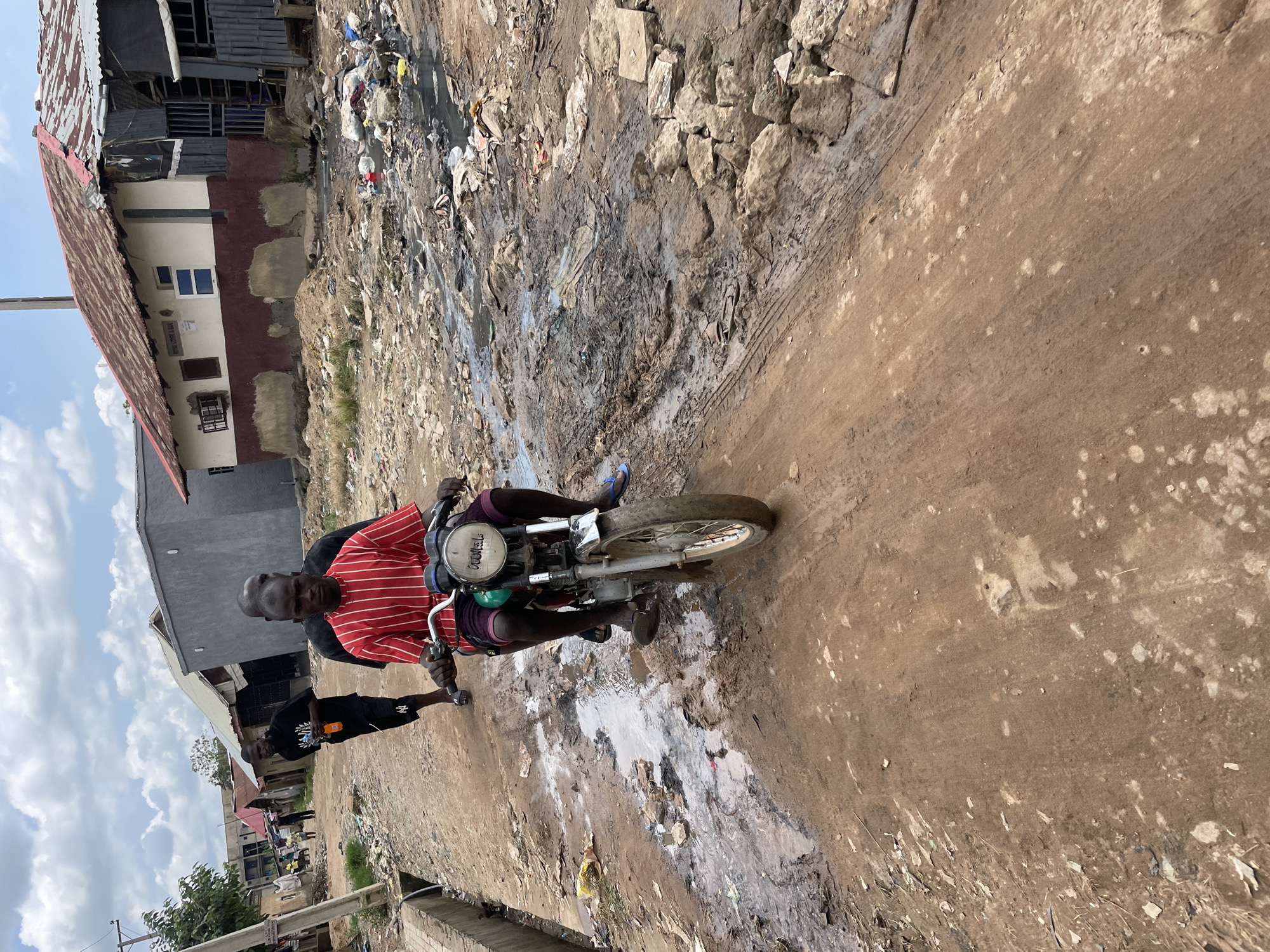 In Abuja, Nigeria, two young men ride a motorbike on a muddy, unpaved road. The driver, wearing a red and white striped t-shirt and dark shorts, is focused on navigating the challenging terrain. Behind him, another young man, dressed in a black t-shirt, watches the road ahead. The environment is characterized by dilapidated buildings, piles of garbage, and stagnant water, suggesting a neglected urban area. The sky is partly cloudy, indicating it might be a humid day. The overall scene depicts a difficult living condition, likely in an informal settlement within Abuja.