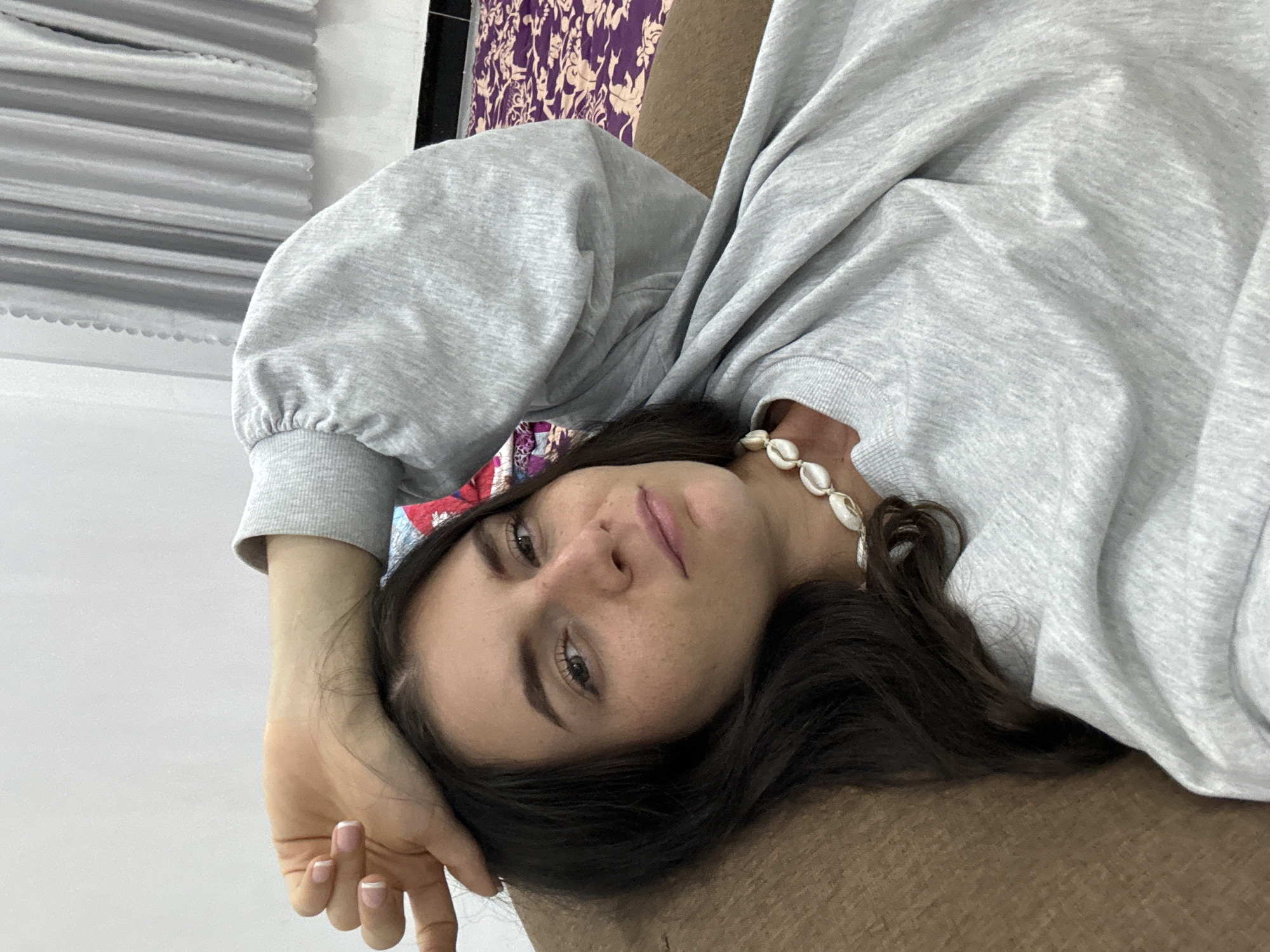 A young woman is captured in a close-up portrait, lying down and looking directly at the camera. She has dark hair and fair skin with visible freckles across her nose and cheeks. She is wearing a casual light grey long-sleeved sweatshirt with gathered sleeves and ribbed cuffs. Around her neck, she sports a distinctive necklace made of large, irregular white, shell-like beads. Her right hand is visible with a French manicure, resting near her head. She appears relaxed, with a calm expression.
The background suggests an indoor setting. To the left, a light-colored, possibly white, wall or textured surface is visible. Behind her head and shoulder, a patterned fabric in purple and cream, resembling a floral or damask design, is seen, likely part of upholstery or a curtain. She is resting on a textured brown surface, such as a sofa or bed. The image is rotated 90 degrees clockwise, depicting her reclining sideways. The lighting is soft and even, indicating an indoor environment during daytime or with good artificial light. There is no visible text in the image. The location context for this scene is Mũi Né, Vietnam.