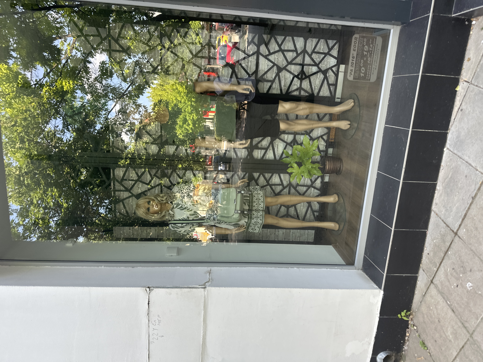 The image shows a storefront window of a shop in Montana, Bulgaria. Two mannequins are displayed inside the window. One is a female mannequin wearing a dress and the other is a male mannequin wearing shorts. A sign displays the business name "PA 2016" EOOD and the working hours. The reflection in the glass shows trees and possibly street scenes, creating a busy, layered effect. A tag is present on the mannequins clothes. Writing is visible on the wall below the window. The sidewalk is visible in the lower right corner of the image.