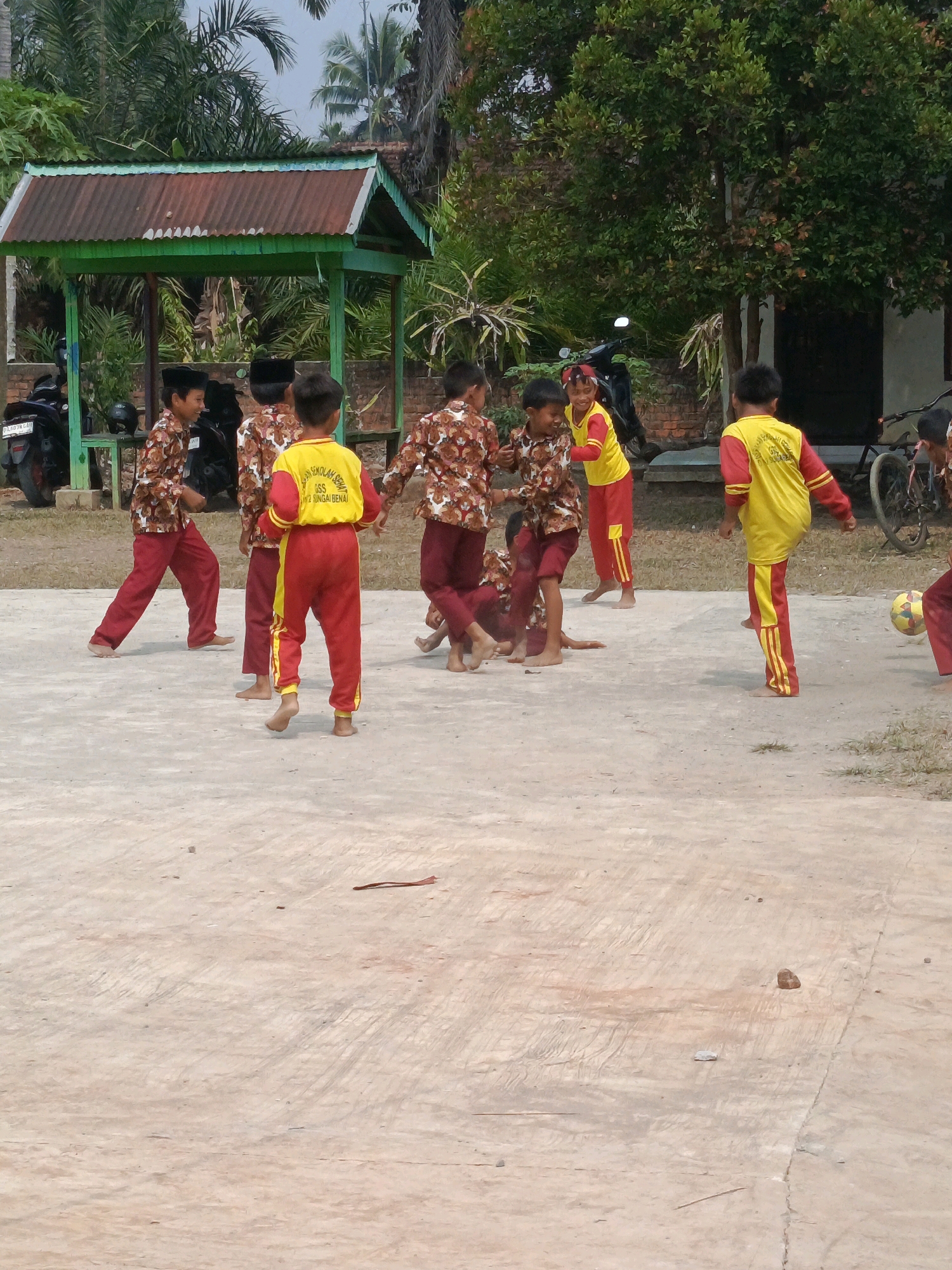 In the city of Megang Sakti IV, Indonesia, a group of young boys, likely students, are engaged in a playful activity on a concrete outdoor court. They are dressed in matching uniforms: some wear brown patterned shirts with red pants, while others sport yellow shirts with red and yellow striped pants. The environment features a rustic, open-air shelter with a corrugated metal roof and green wooden posts, surrounded by lush green vegetation and palm trees. In the background, a building and a bicycle are visible. The boys appear to be playing with a yellow and black soccer ball, with some actively kicking it and others observing. The scene is bathed in bright daylight, suggesting an outdoor educational or recreational setting.
