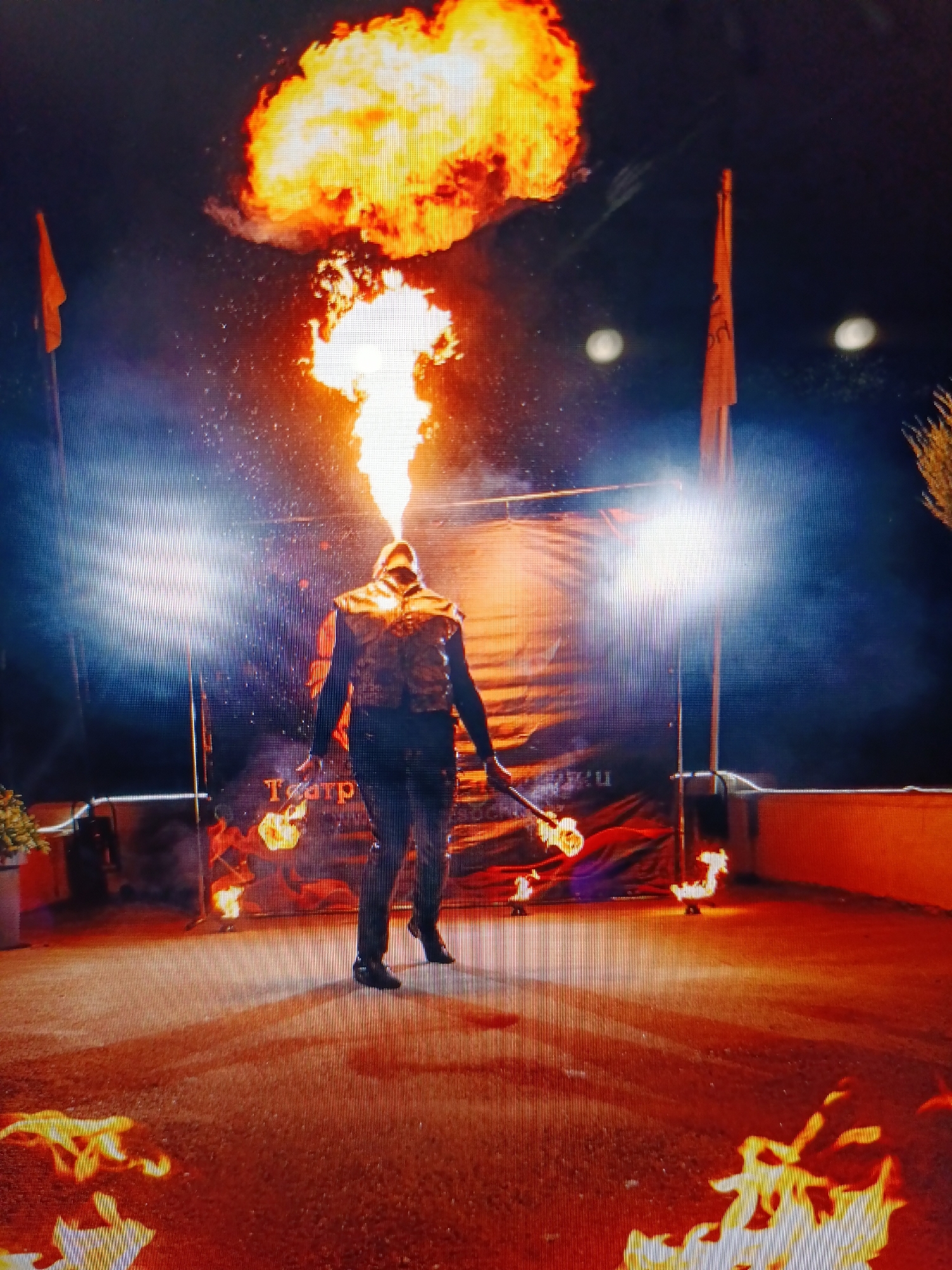 A fire performer is pictured in the foreground, holding a flaming torch in their right hand and expelling a large plume of fire from their mouth. They are wearing a dark, textured jacket and dark pants, with black shoes. The performer is standing on a reddish-brown, granular surface with scattered flames. In the background, two tall orange flags on poles stand to the left and right of the performer. Behind the performer, a large black banner with an orange design is visible, along with two bright spotlights. The sky is dark. The scene appears to be outdoors at night, possibly at an event or performance.