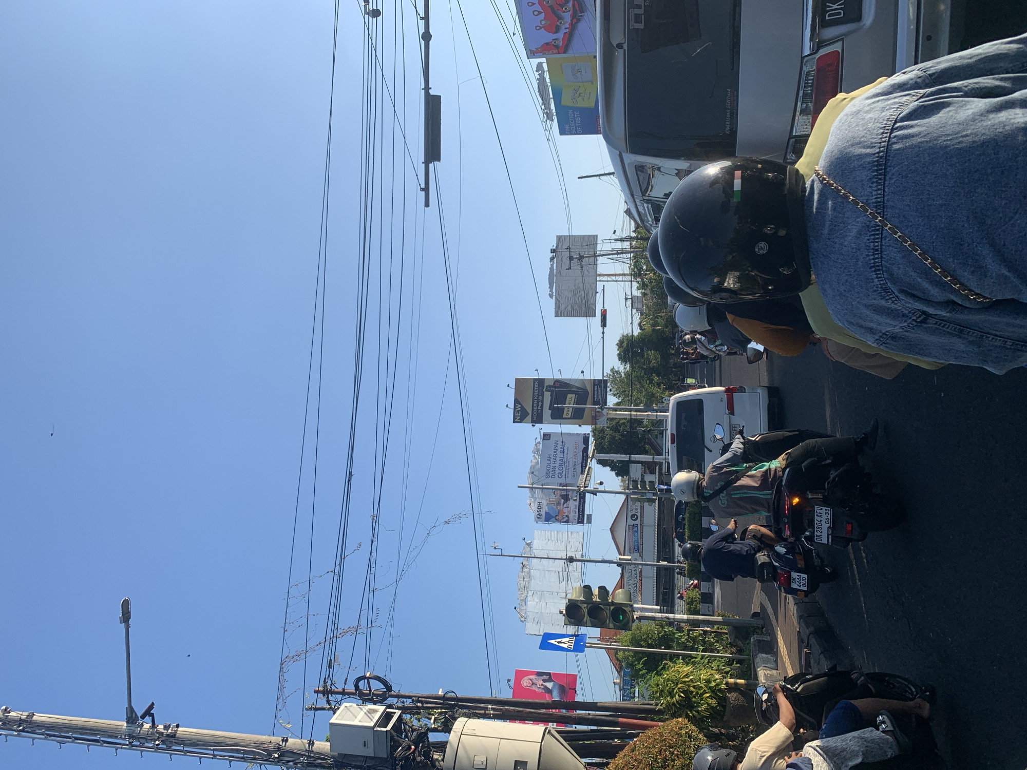 The image captures a street scene in Denpasar Barat, Indonesia, during daylight hours. The perspective is from the back of a motorcycle, looking forward at a traffic intersection. In the foreground, the backs of several individuals on motorcycles are visible, wearing helmets. One person in the immediate foreground wears a blue denim jacket and a black helmet with an Italian flag sticker.
The street is paved, with a traffic light showing green for the direction of travel. Several motorcycles and a white van are stopped or moving slowly in traffic. Billboards are erected on poles along the side of the road, some displaying advertisements and text, including one with the words "SDH SEKOLAH IMPIAN HARAPAN GLOBAL BALI" and "OPENING JULY 2021." Overhead, a complex network of electrical wires and poles crisscrosses the sky, which is clear and blue. Vegetation, including trees and bushes, can be seen along the side of the road.