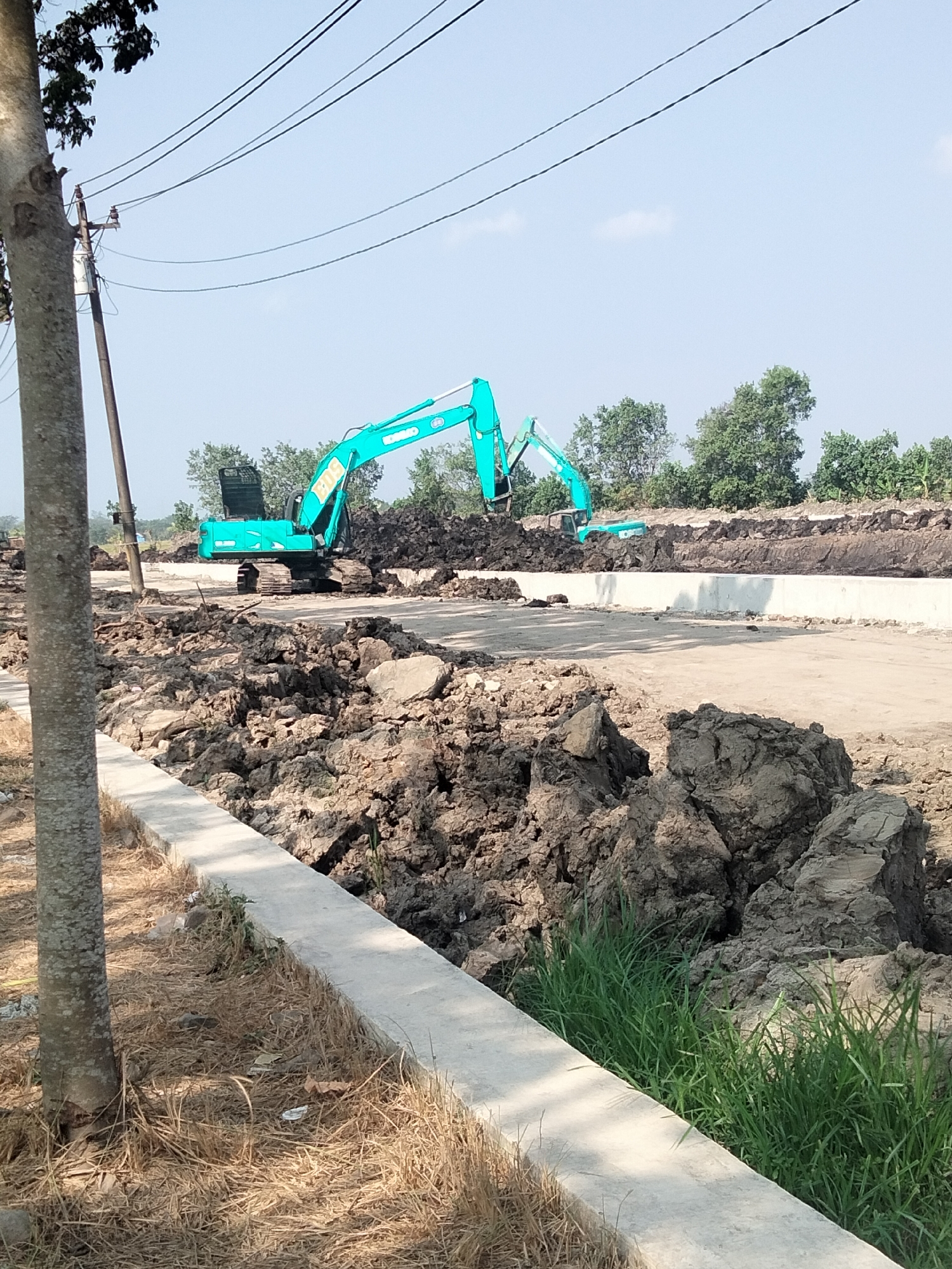 In Sikampuh, Indonesia, an outdoor construction scene is captured under a clear sky. Two teal excavators are prominently positioned. One is in the middle ground, operating near a large mound of dark soil, its arm extended. Another, partially visible, is further in the background. The terrain consists of disturbed earth, piles of soil, and a concrete structure running diagonally across the scene. A lone tree stands on the left, its trunk in the foreground. Overhead, electrical wires stretch across the frame. The environment appears to be a developing area, possibly for infrastructure or building.