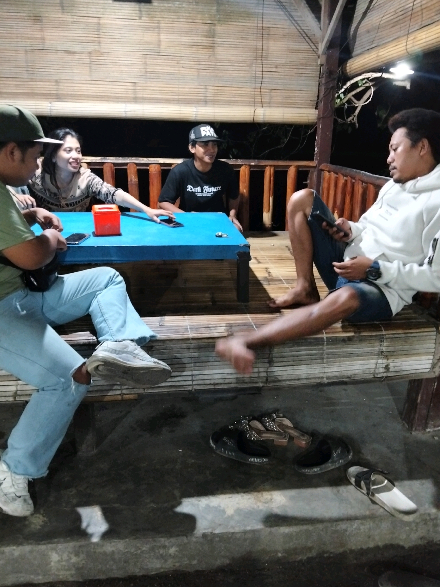 Four individuals are gathered at a wooden, open-air structure, likely a gazebo or a patio, with a bamboo-paneled ceiling and wooden railings. The ground is concrete, and a low wooden bench is situated in the foreground. A blue rectangular table is the central focus of the group.
On the left, a person wearing a green t-shirt, a baseball cap, and light blue ripped jeans sits with their legs extended. They appear to be holding a mobile phone. Next to them, a woman with long dark hair smiles and leans forward, her hands resting on the table. She wears a patterned sweater.
To the right of the woman, a person wearing a black t-shirt with white lettering and a black baseball cap is seated. Their hands are on the table, and they are looking forward.
On the far right, a person with dark curly hair, wearing a white hooded sweatshirt and dark shorts, is seated on the wooden bench, looking down at a mobile phone in their hands.
On the blue table, there is a red tissue box and at least two mobile phones. In the foreground, on the concrete floor, several pairs of sandals are scattered. The lighting suggests it is evening or night. The scene appears to be a casual gathering in an outdoor, possibly rural or semi-rural, setting in Indonesia.
