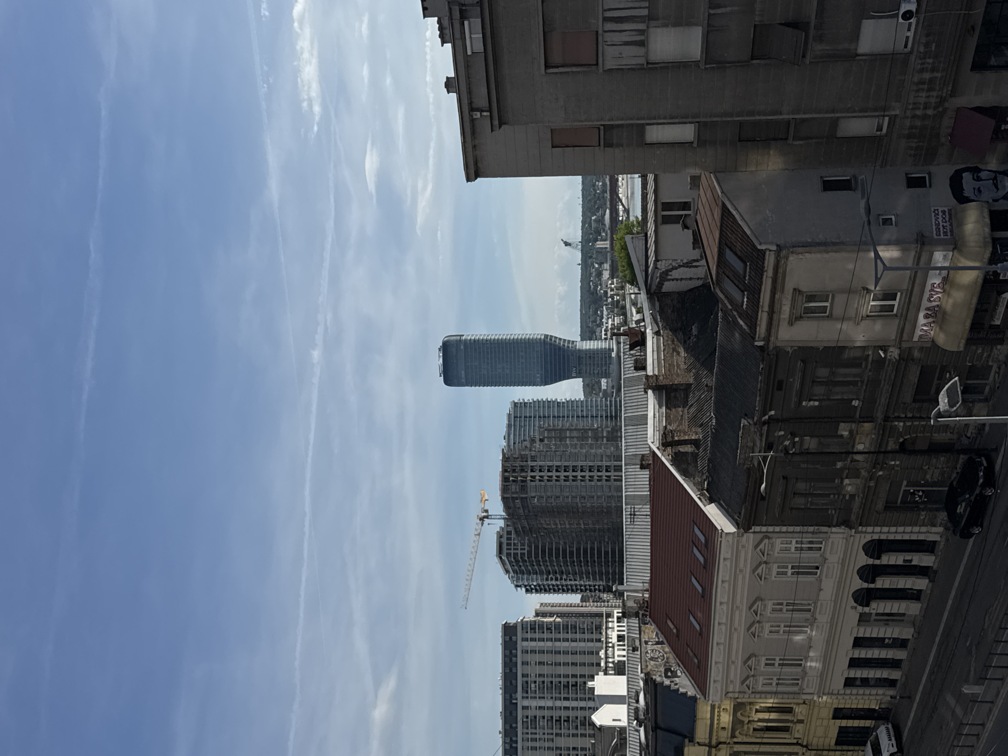 The image shows a view of Belgrade, Serbia, dominated by modern skyscrapers under construction and older buildings in the foreground. In the center, a distinctive curved skyscraper stands out, while a construction crane indicates ongoing building activity. The sky is blue with streaks of clouds. The foreground features a mix of older, detailed building facades, some with red tiled roofs. A street with parked cars runs along the bottom right of the frame. Overall, the scene depicts urban development juxtaposed with older architecture in Belgrade.