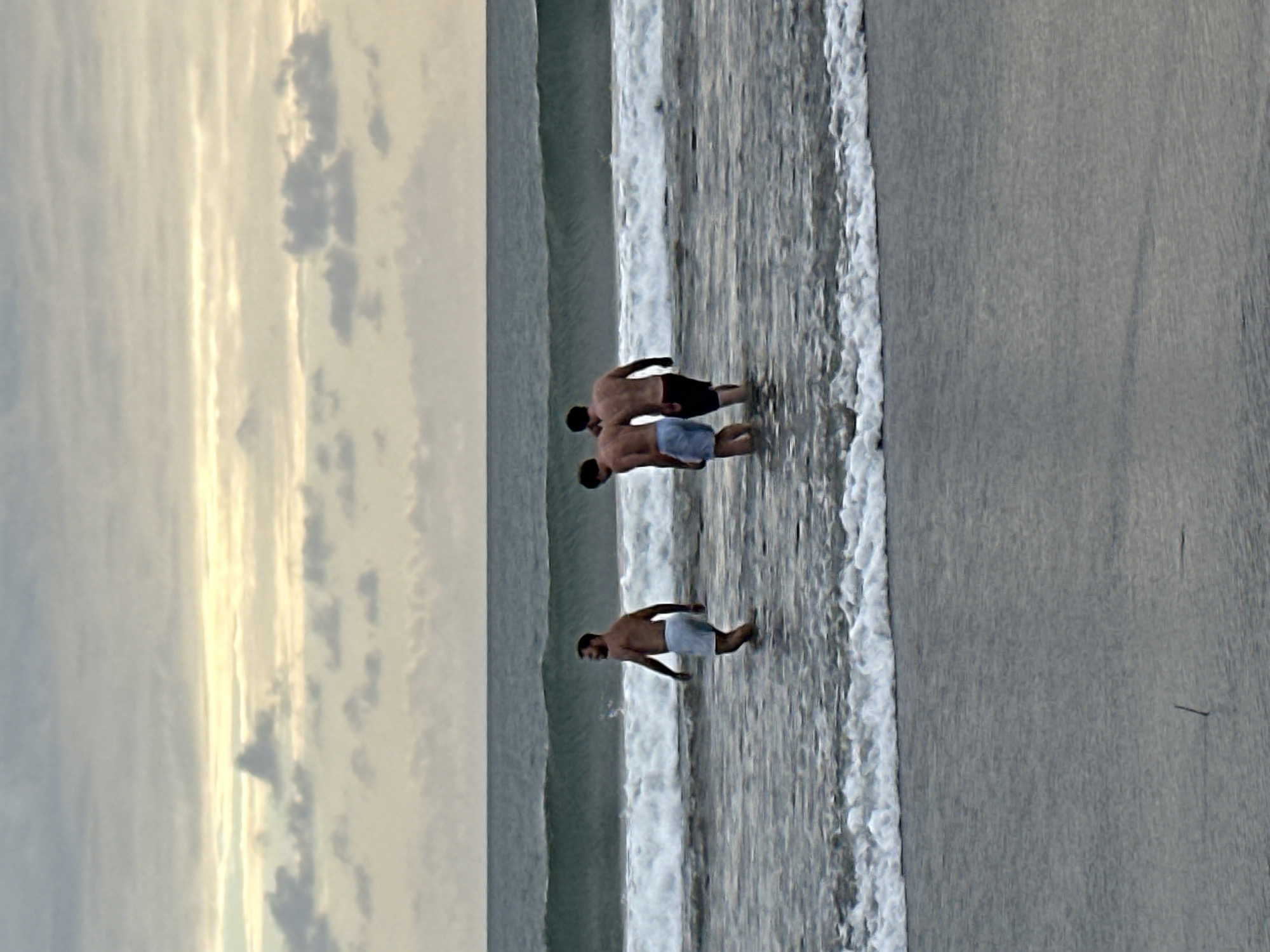 Three shirtless men are standing in the ocean in North Kuta, Indonesia. The man on the left is wearing light blue shorts and is facing to the left. The man in the middle is wearing blue shorts and is facing away from the camera. The man on the right is wearing dark shorts and is facing away from the camera. There are waves in the ocean and clouds in the sky. The scene appears to be during the day.