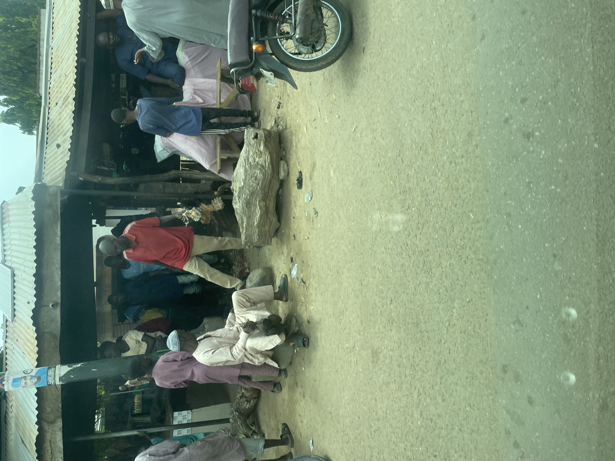 In Chabal, Nigeria, a busy street scene unfolds in front of a corrugated metal-roofed stall. Several individuals are gathered, some in discussion and others engaged in various activities. A man in a red shirt stands prominently, holding a cluster of dead chickens. Next to him, a man in a light-colored robe is seated, holding another chicken. A motorcycle is parked to the right, its rear wheel visible. In the background, more people mill about, and a large piece of wood rests on the ground. A poster of a man in blue is affixed to a pole, suggesting a political or public figure. The ground is dusty and uneven, indicative of an outdoor market or roadside setting.