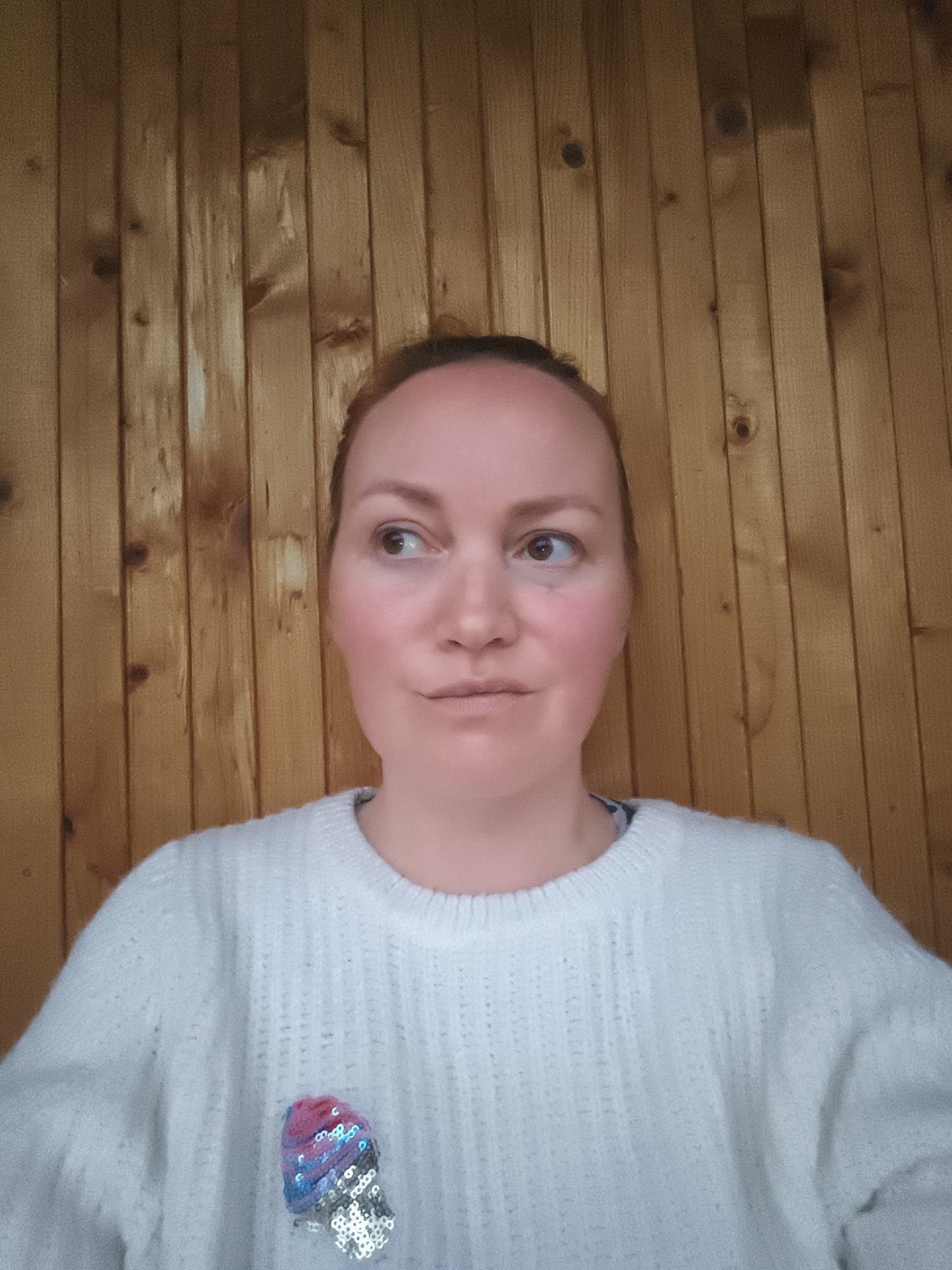 A fair-skinned individual, likely female, with reddish hair pulled back, is positioned centrally against a vertical wooden plank wall. The person's brown eyes gaze upwards and to the viewer's left, with a neutral expression on the lips. They are wearing a light-colored, possibly off-white, textured knit sweater. An embellished patch or brooch resembling an ice cream cone, composed of pink, blue, and silver sequins, is visible on the left chest area of the sweater. The wooden background features prominent grain patterns and knots, illuminated by soft, natural light, in the city of Unknown, Unknown.