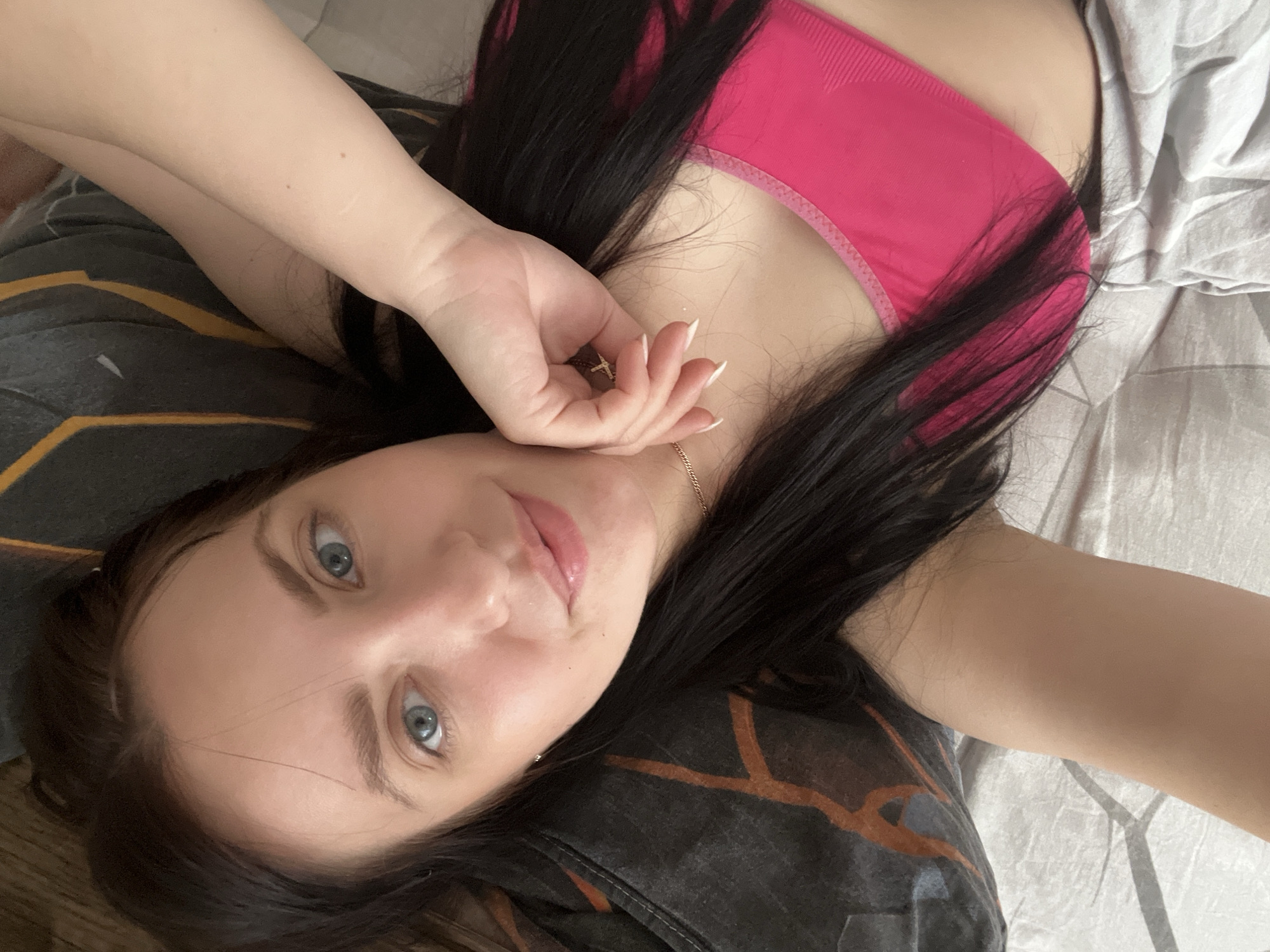 A woman with long, dark hair, fair skin, and blue eyes is lying on what appears to be a bed. She is wearing a pink top and appears to be indoors. Her hand is near her face. Part of a patterned pillow is visible under her head, and light-colored bed sheets are to the side. There is no visible information to determine the location as Марковский, Russia.