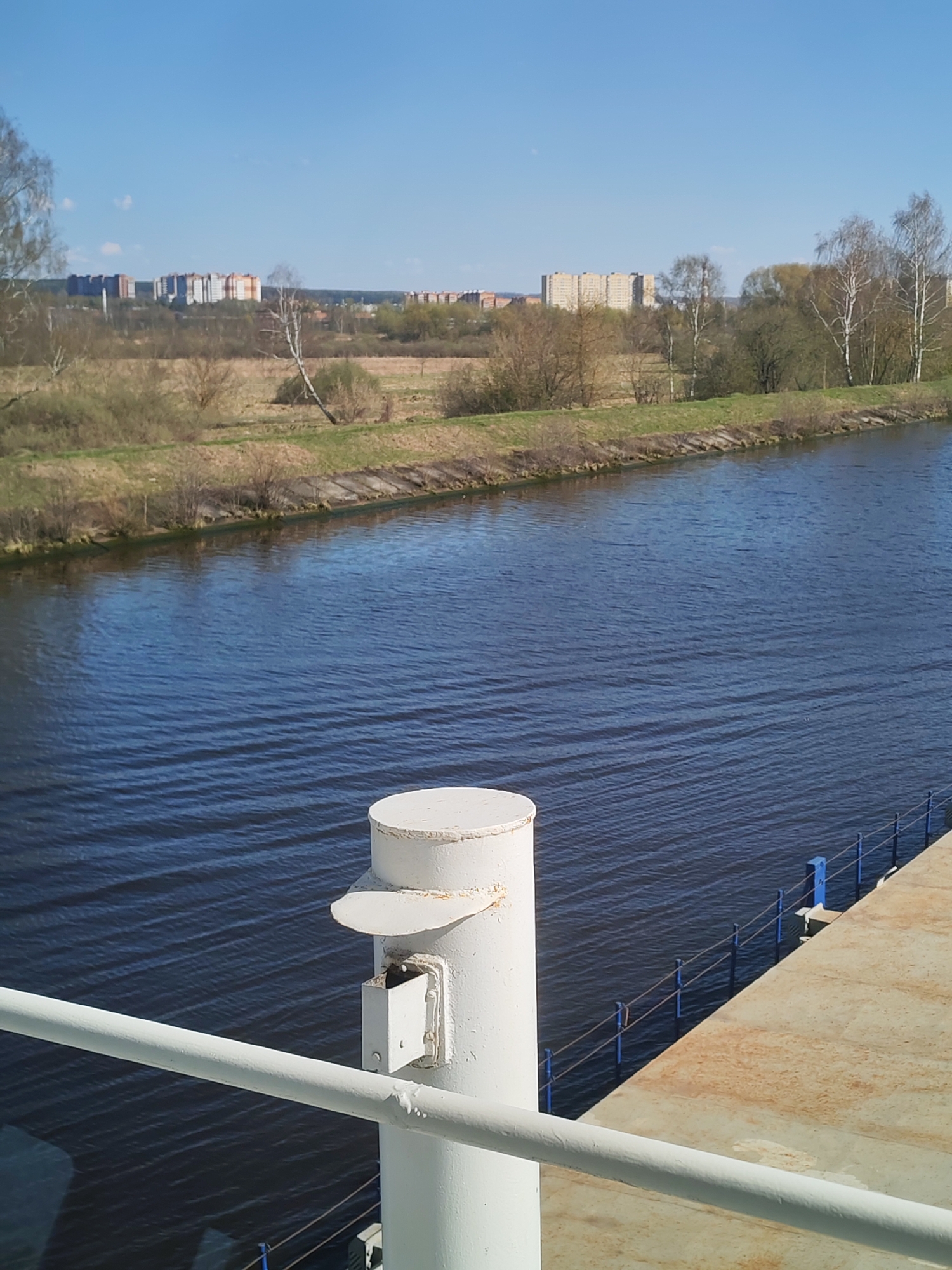 The image depicts a waterway, possibly a canal or river, with urban structures visible in the distance. The foreground features components of a ship or dock, including a white cylindrical pillar and railing. The waterway is lined by vegetated banks. The sky is blue and cloudless. The location is Unknown, Unknown.