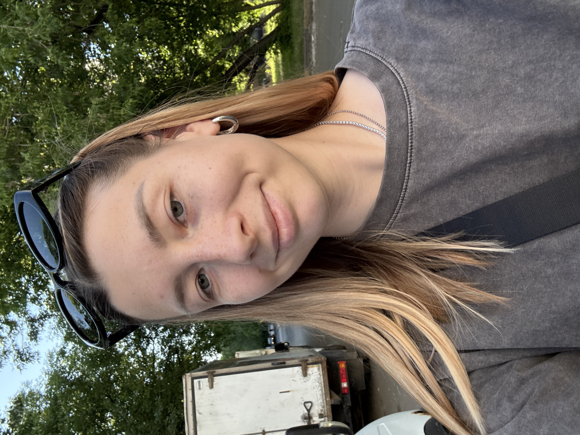 A young woman is depicted in a close-up, head-and-shoulders portrait, situated outdoors in what is identified as Moscow, Russia. She has long, straight, light-brown hair with blonde highlights and fair skin with visible freckles. Her eyes are light-colored, and she exhibits a subtle, closed-lip smile. She wears black sunglasses pushed up onto her head, silver-colored hoop earrings in both ears, and a two-strand silver chain necklace. Her attire is a dark grey, stone-washed crew-neck top, with a dark fabric strap visible diagonally across her chest. The background shows a sunny daytime environment with a clear blue sky partially visible through dense green tree foliage. To the left, a white utility truck with rear doors and taillights is parked on a paved surface, with another similar truck further down the street. A portion of a white vehicle is visible in the lower left foreground.