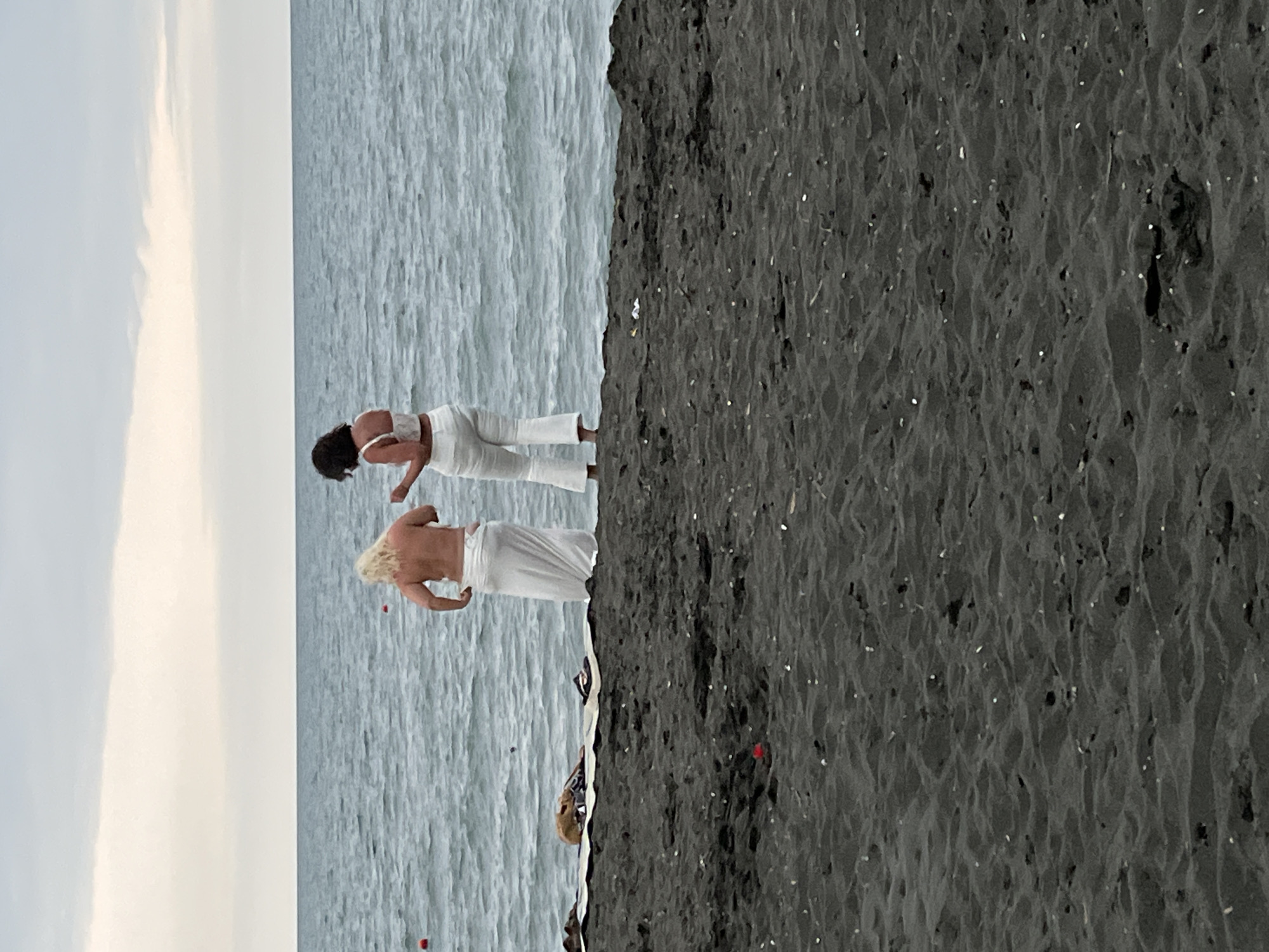 Two individuals stand on a black sand beach, facing the sea. The person on the left has blonde hair and is wearing a white wrap around their lower body, exposing their bare chest. The person on the right has dark hair and is dressed in a white crop top and white flared pants. The sea is calm with visible ripples and a few red buoys in the distance. The sky is overcast. The beach is covered in dark sand with small white shells or pebbles scattered across it. To the left of the two individuals, a folded blanket or towel with a brown object on top is visible. This scene is consistent with Ureki, Georgia, a location known for its black magnetic sand beaches.